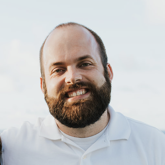 Headshot of a smiling man with a beard and short hair, wearing a white collared shirt, outdoors with a cloudy sky in the background.