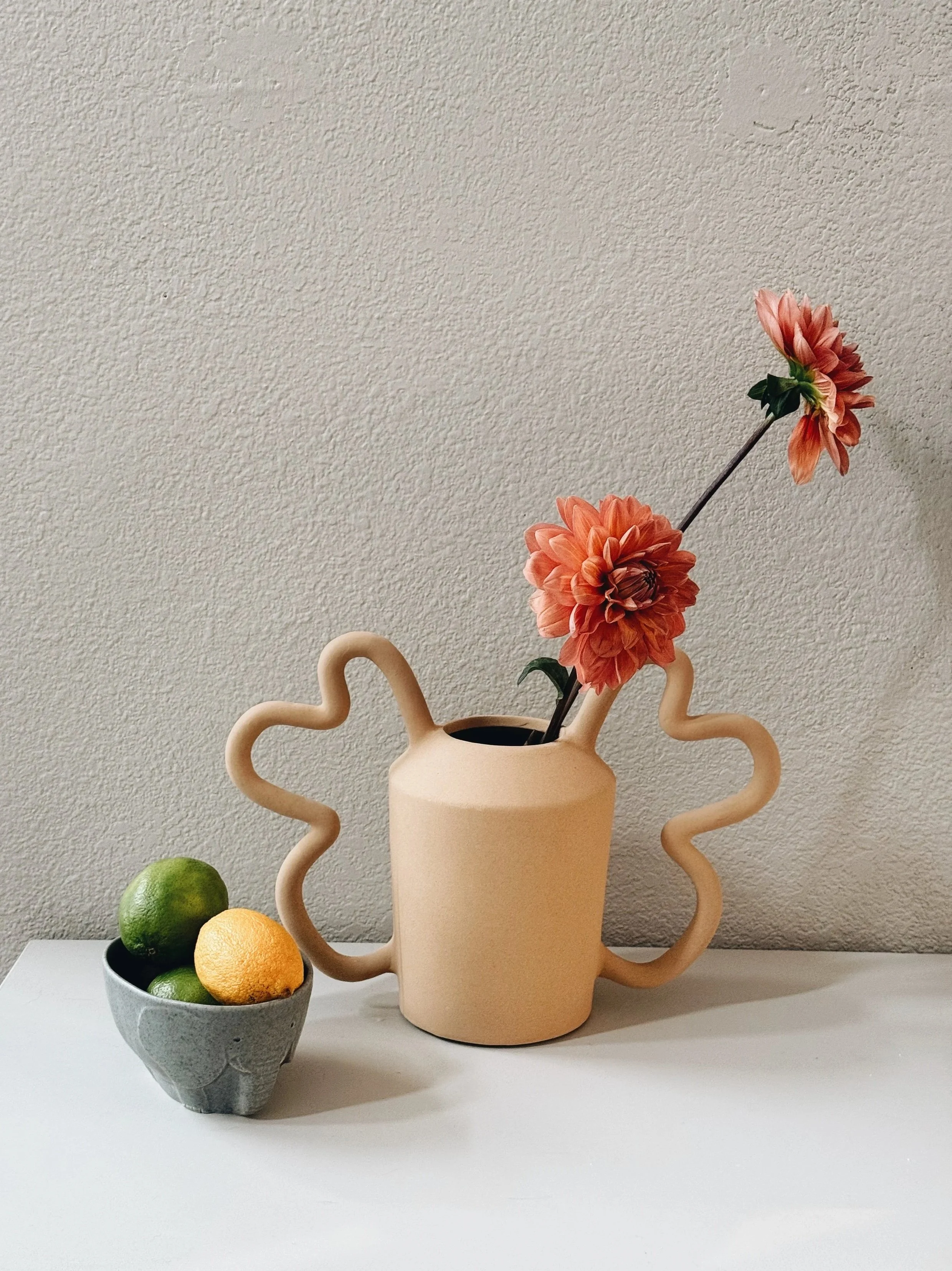 A beige vase with wavy handles containing two peach-colored flowers on a beige wall background. A bowl of green lemons and yellow citruses on a white surface in front of the vase.