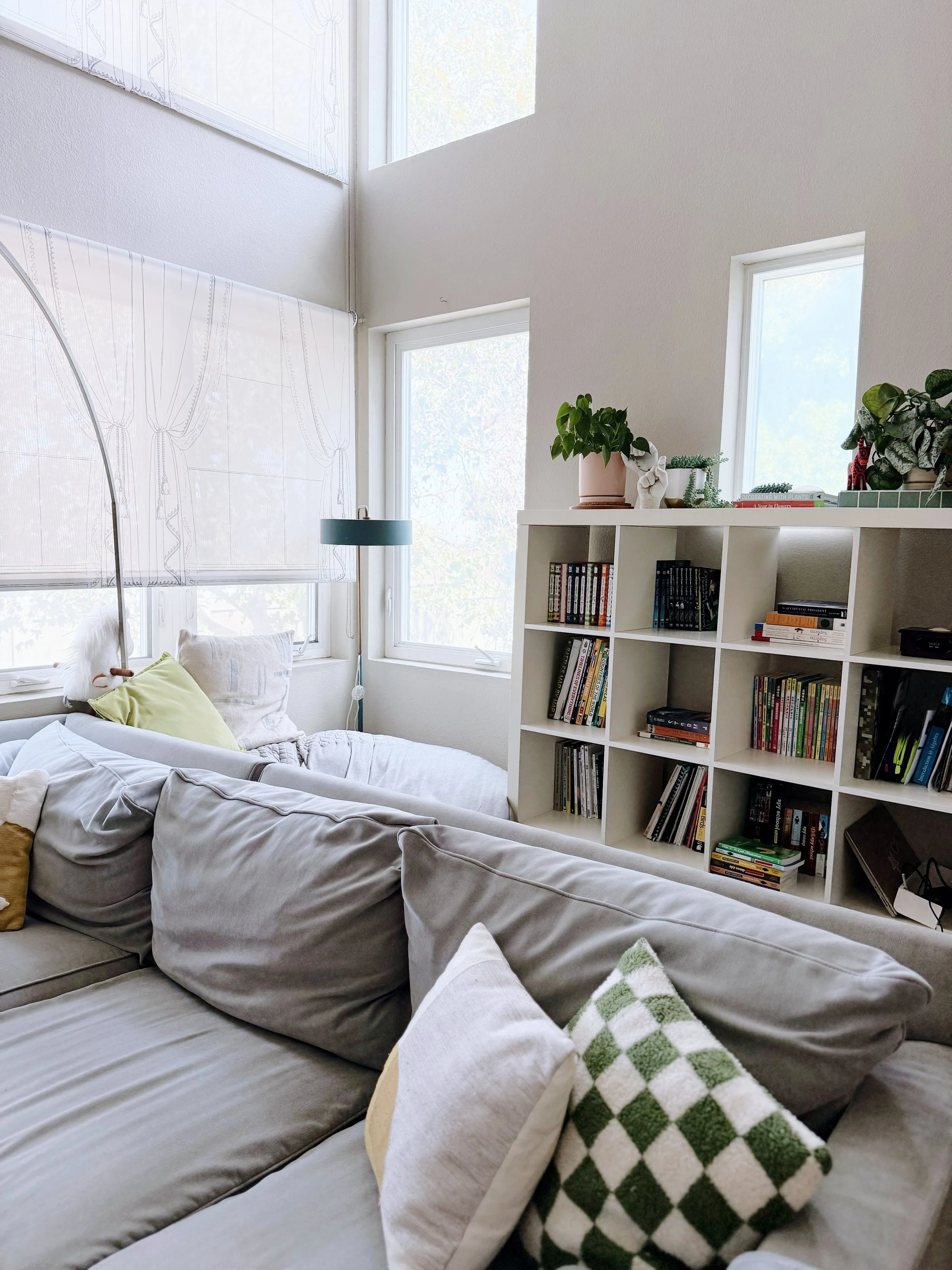 Living room with a beige sofa, throw pillows, a white bookshelf filled with books and decorative plants, large windows with white window treatment, and a white wall.