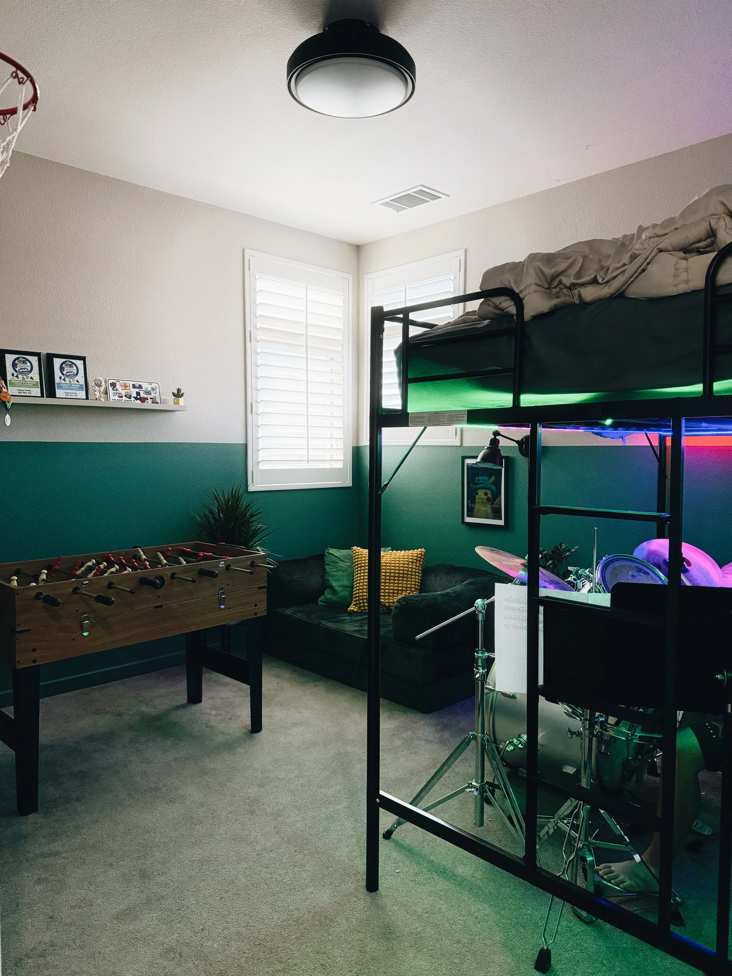 A room with a bunk bed, a foosball table, a couch with pillows, framed photos and decor on a wall shelf, a small plant, and a drum set with LED lights. There are windows with white shutters and a black ceiling light.