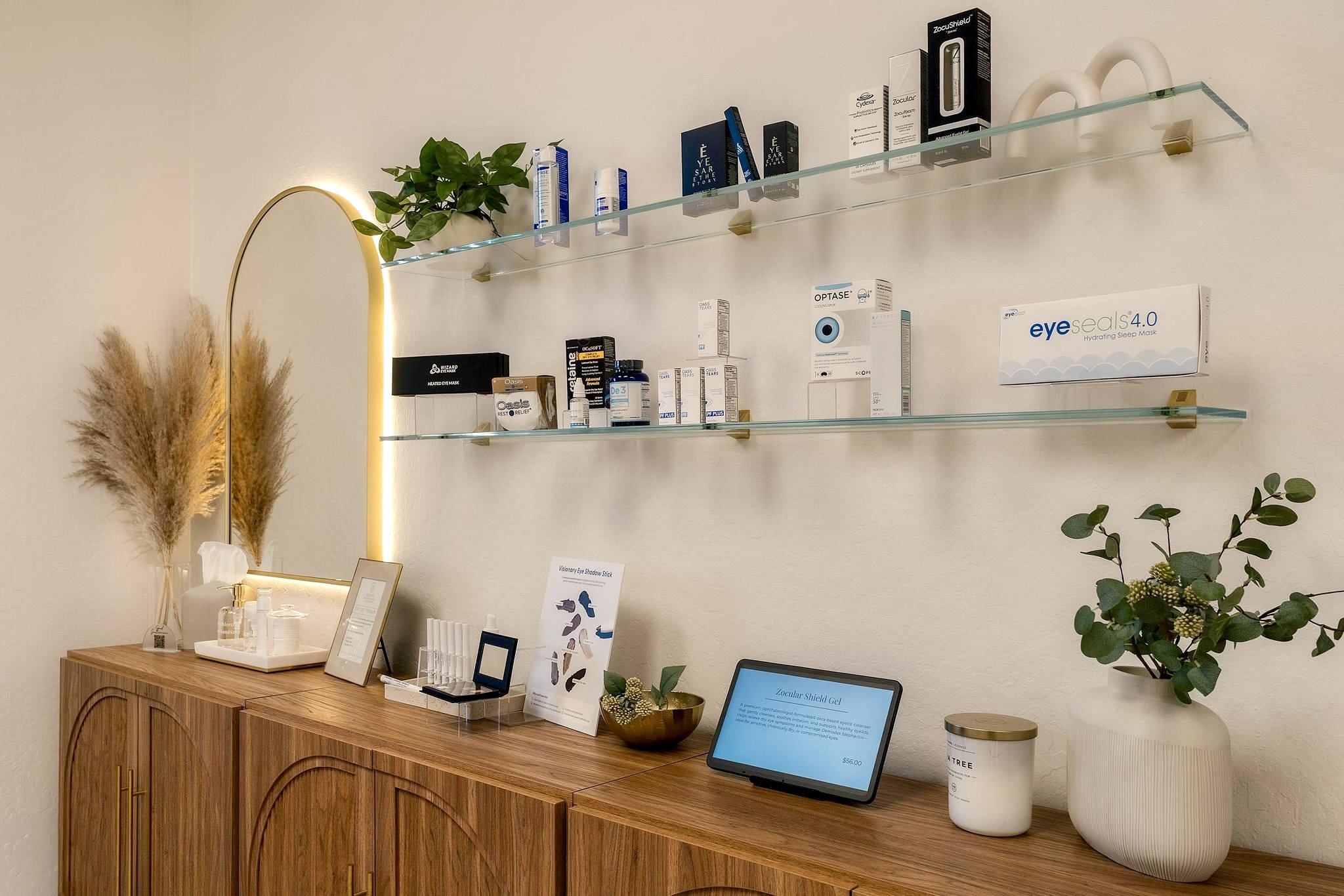 Wooden cabinet with skincare and eye care products, a mirror with a plant to the left, and a digital screen displaying eye masks on the wall.