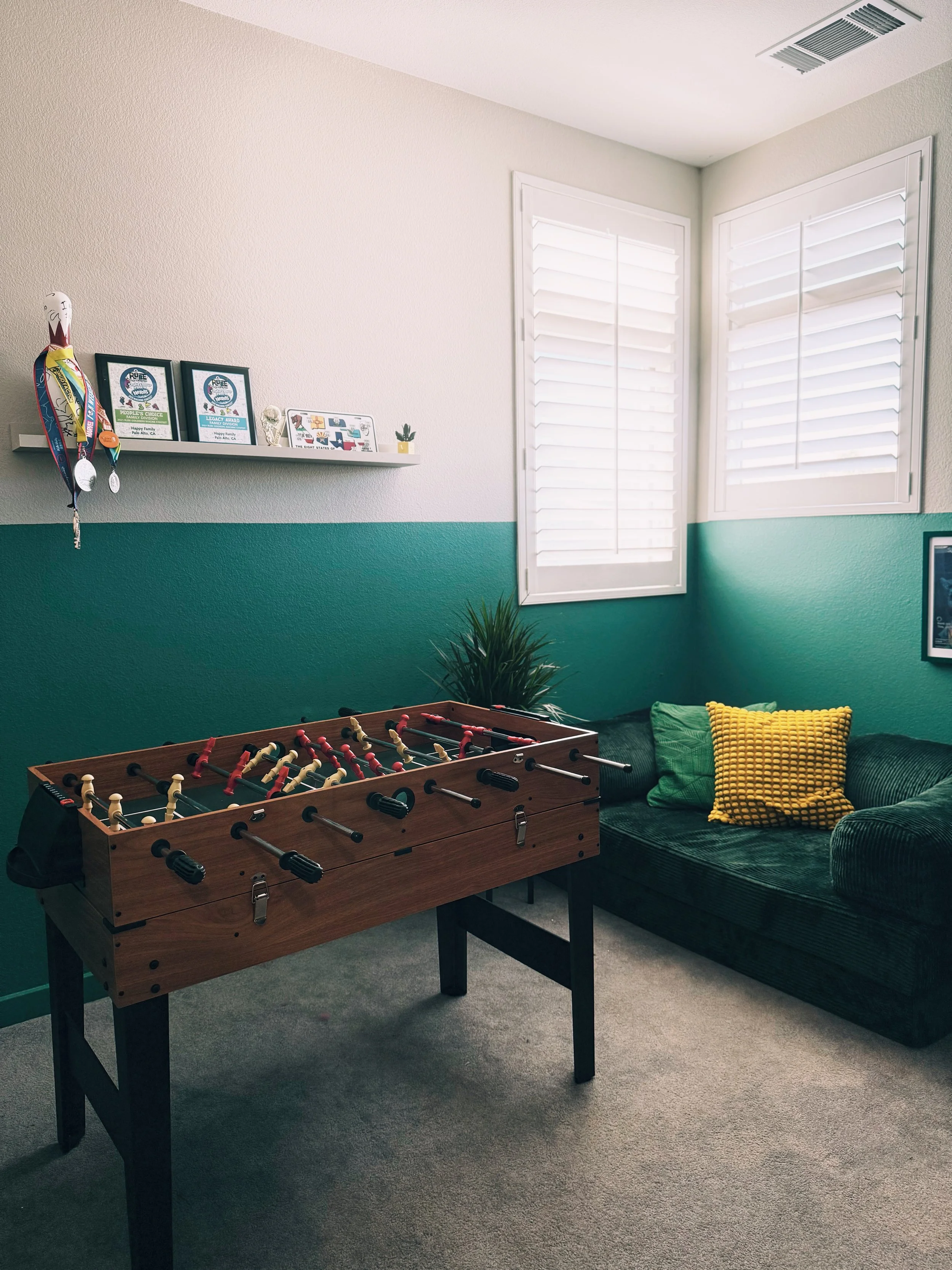 A room with a foosball table, a green couch with a yellow pillow, and a wall shelf with awards and decorations. There are two windows with white shutters and a small plant near the windows.
