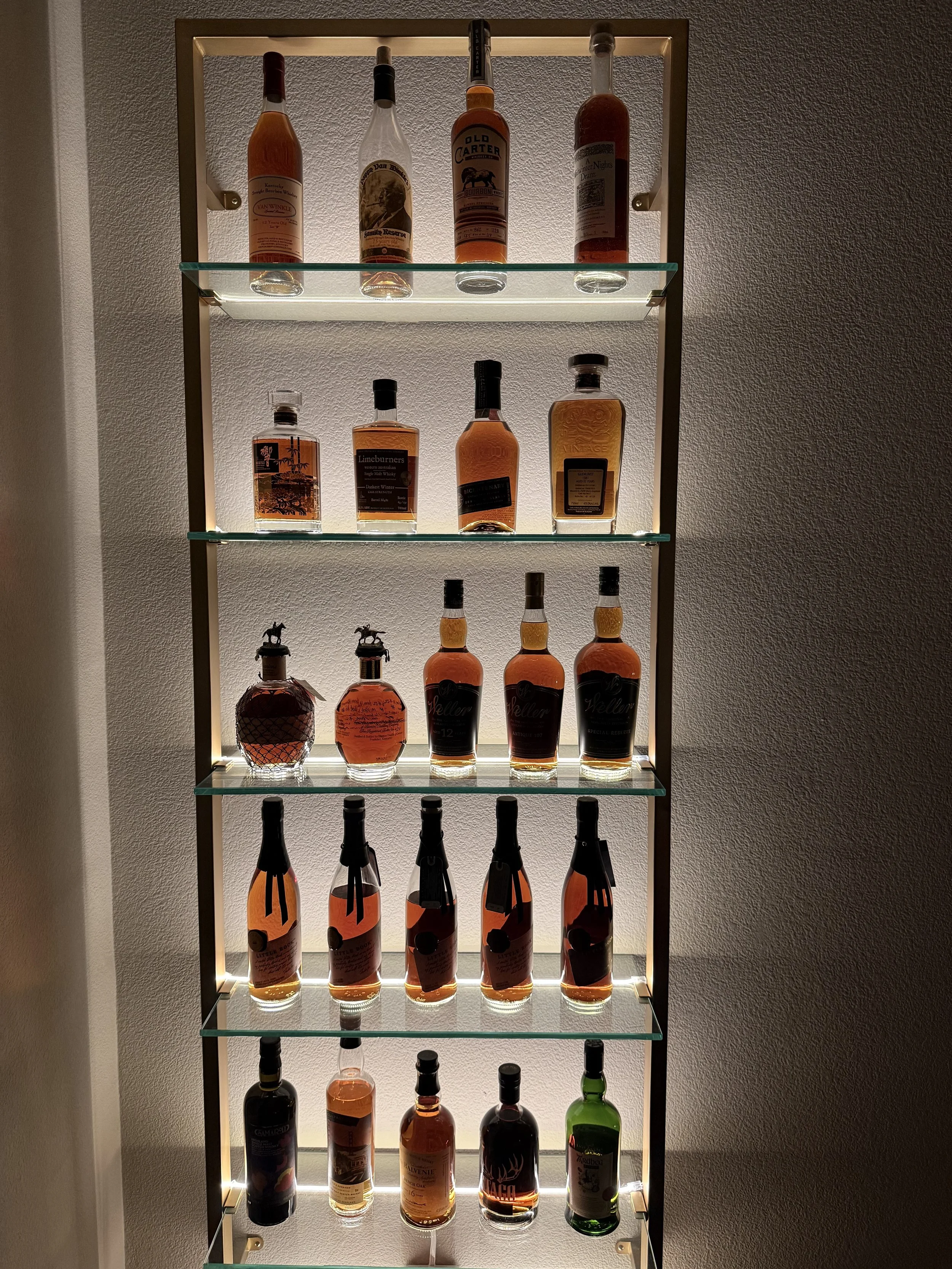 A display case with multiple bottles of liquor on glass shelves, backlit with warm lighting, against a textured wall.