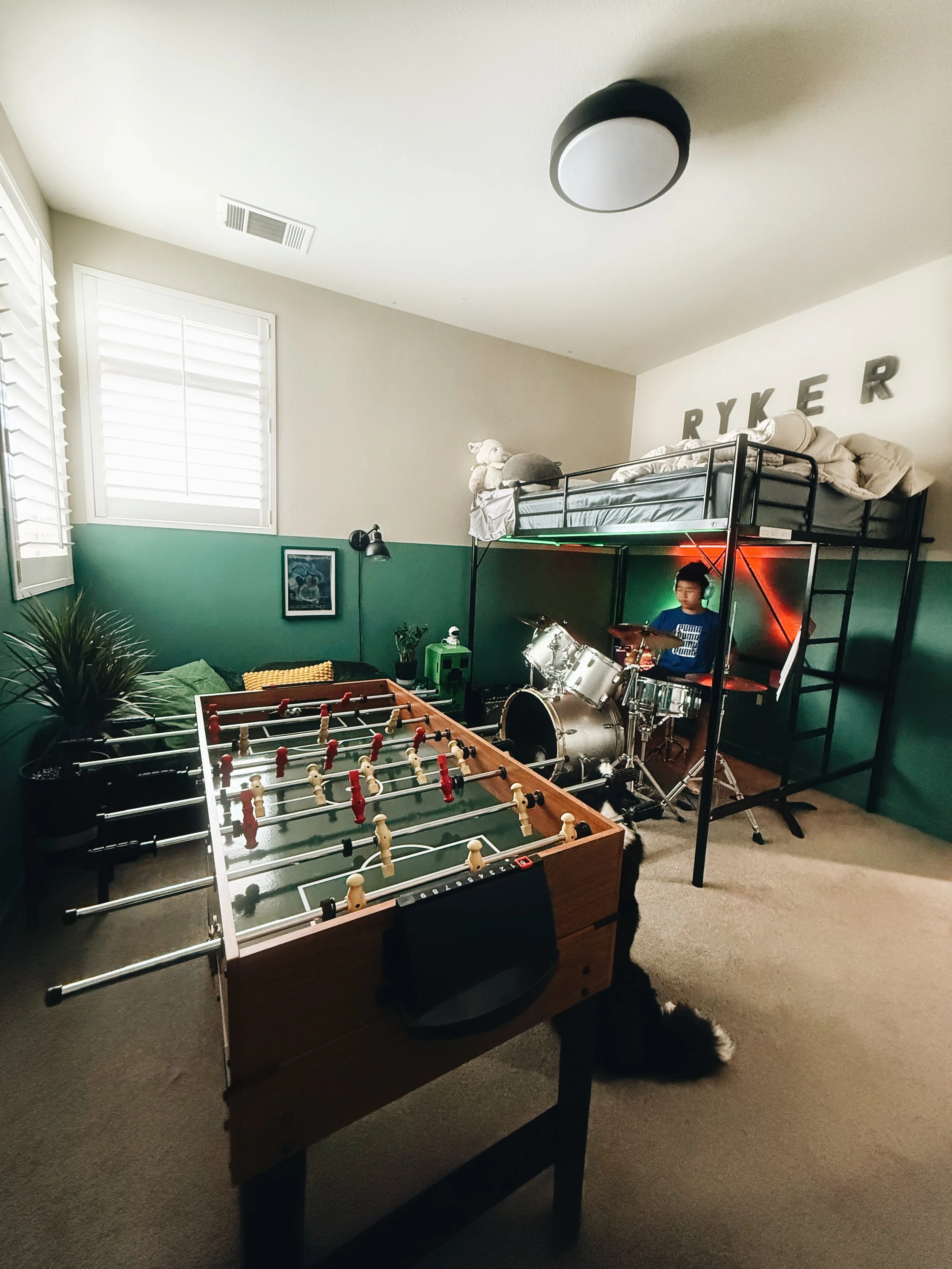 A bedroom with a foosball table in the foreground, a drum set, a bunk bed with stuffed animals and pillows, a window with white shutters, and the name RYKER on the wall above the bed.