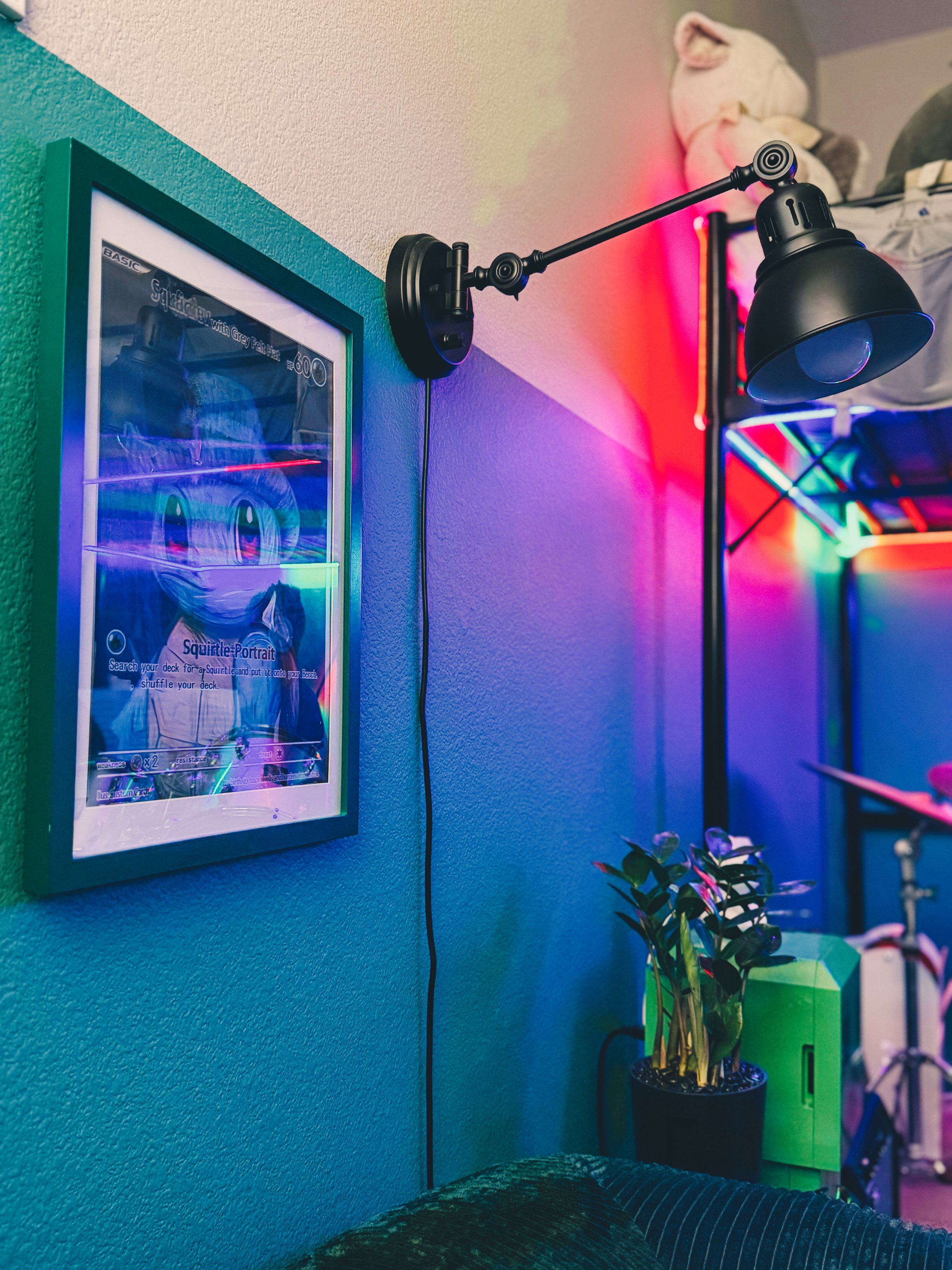 A black wall-mounted lamp with an adjustable arm illuminating a colorful room with neon and LED lights. A framed poster is on the turquoise wall to the left, and a potted plant is on a green cabinet near the bottom right. A stuffed animal is on a she