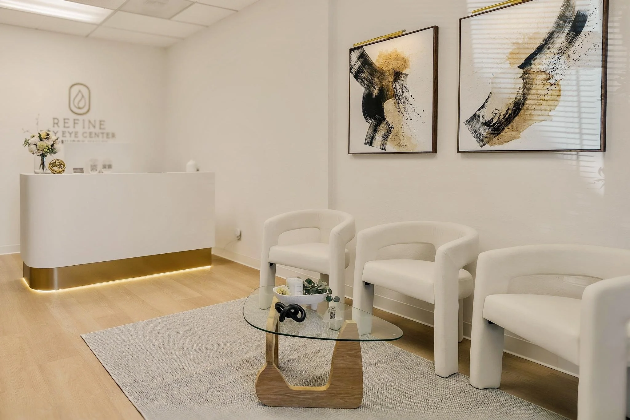 Waiting room of Enhance Eye Center with three white chairs, a glass coffee table with skincare tools, and three abstract paintings on the wall.