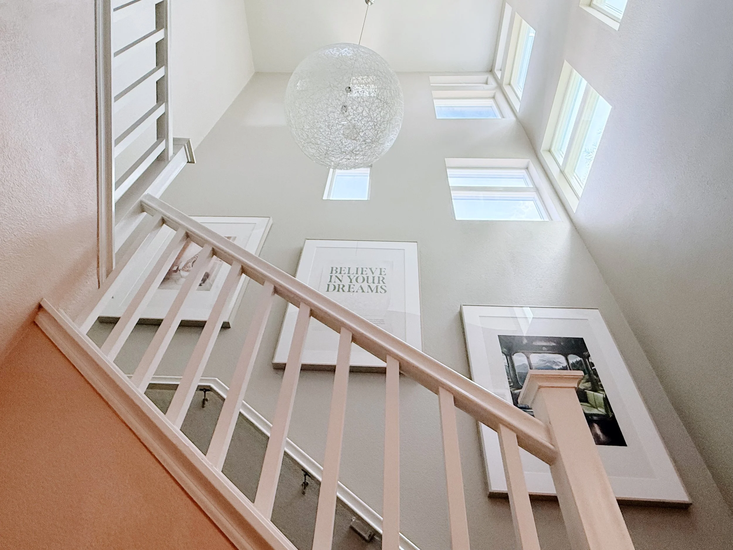 The photo depicts the interior of a house showing a staircase leading up to a second floor with a high ceiling and large windows allowing sunlight to brighten the space. There’s a framed quote on the wall that reads, 'BELIEVE IN YOUR DREAMS,' and a d