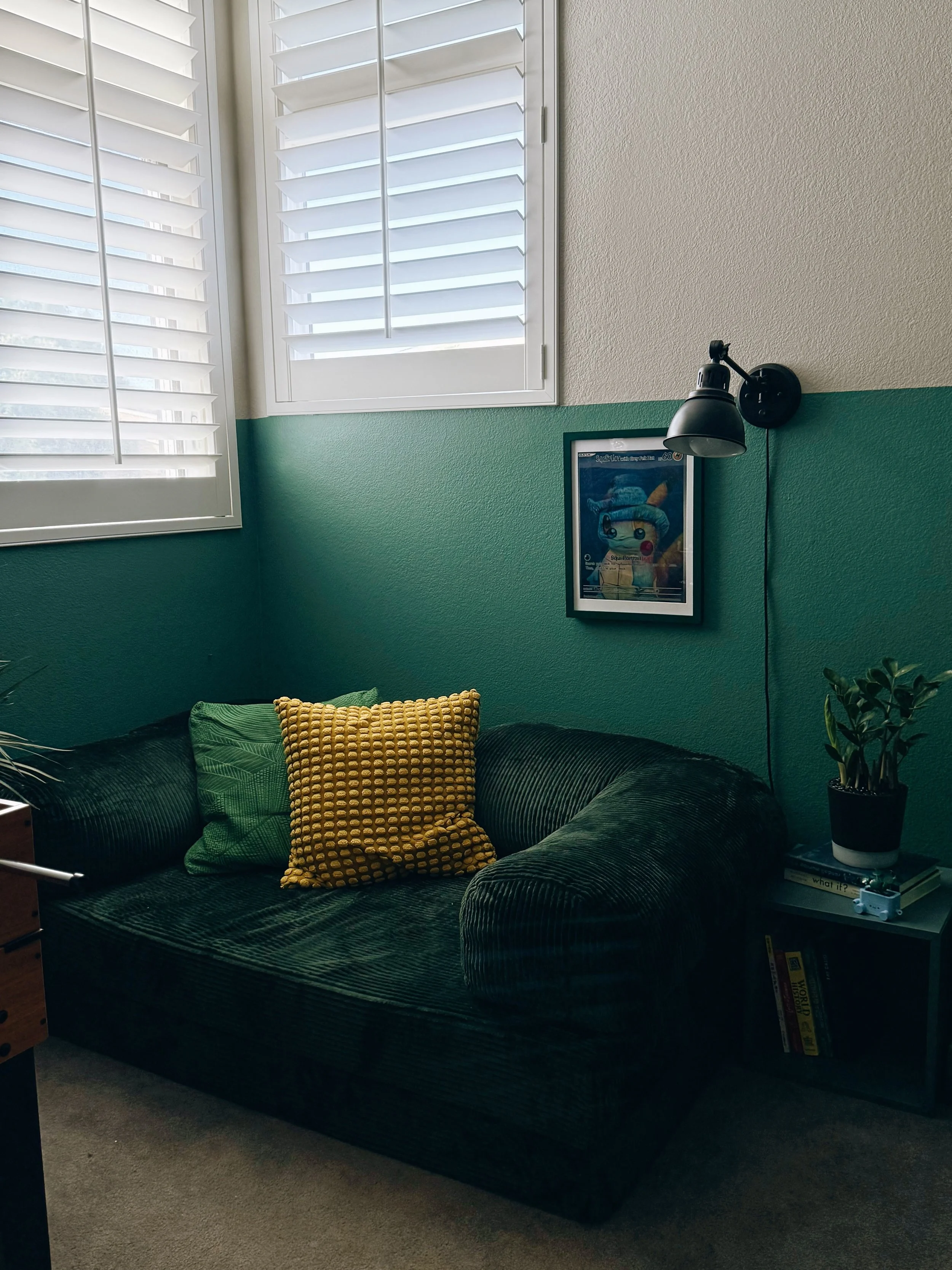 A living room corner with a dark green velvet couch, a yellow textured pillow, a framed picture of Pikachu from Pokémon, a black wall-mounted lamp, a side table with a potted plant and books, and windows with white plantation shutters.
