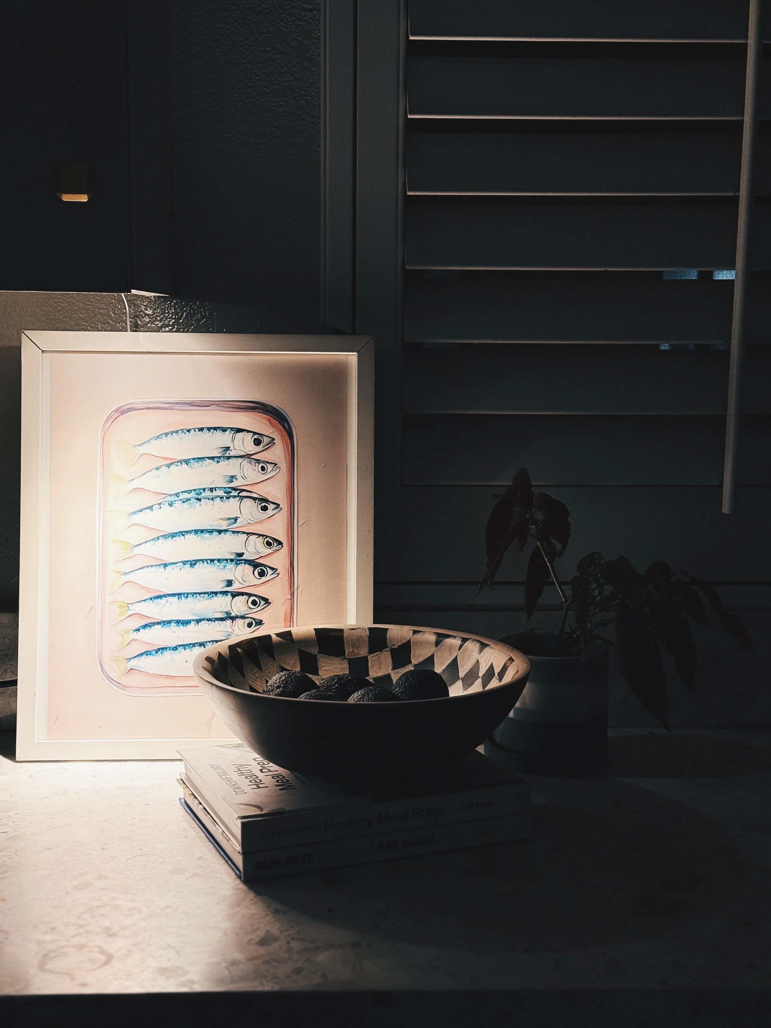A framed illustration of ten fish with blue and silver scales on a kitchen counter. A black and beige patterned bowl filled with dark round objects is in front of the illustration, with a stack of books underneath it. The background includes a black 
