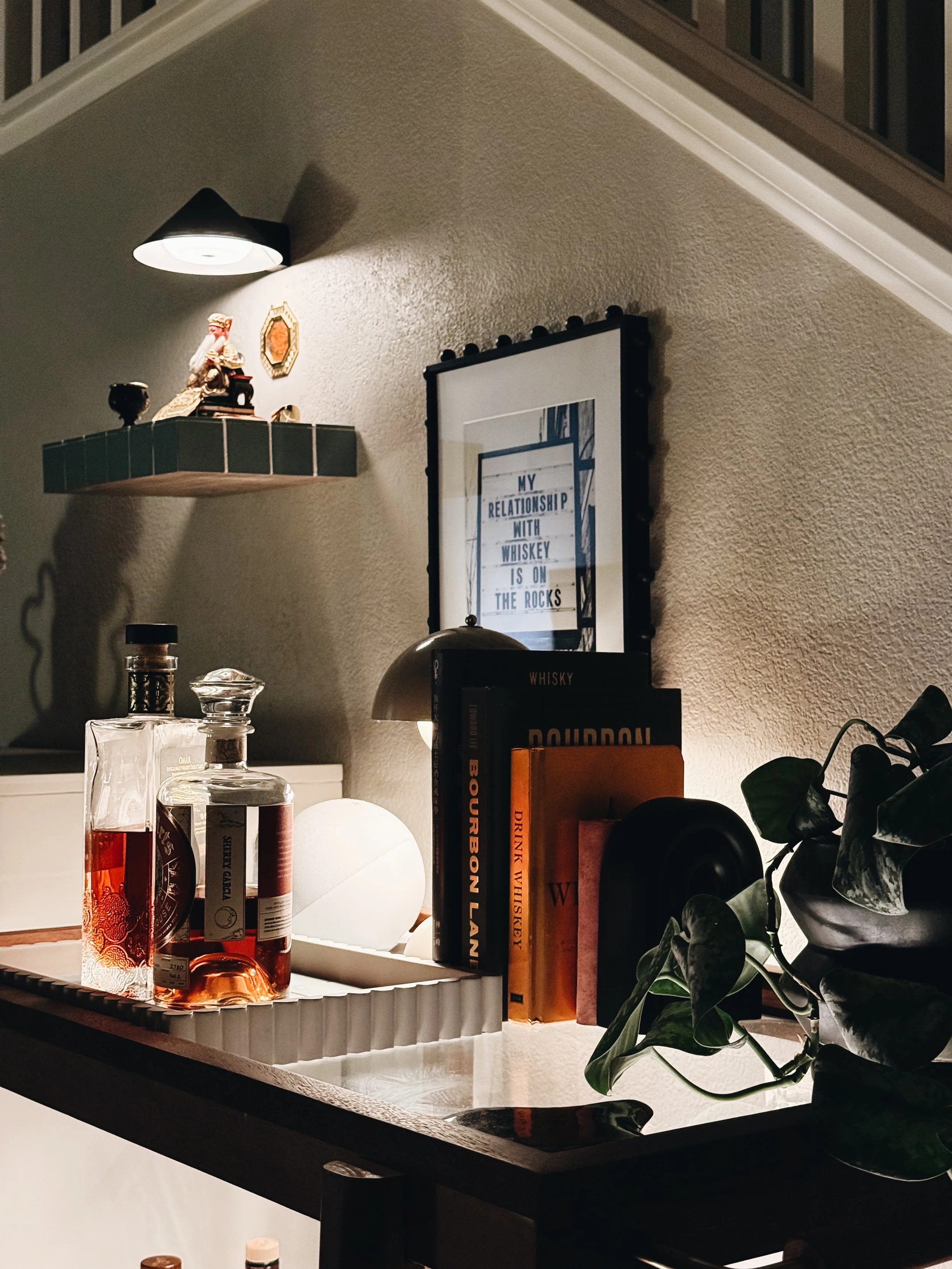 A bar cart with bottles of alcohol, a decorative figure, a lamp, books, and a plant, against a textured beige wall with a wall light and framed artwork.