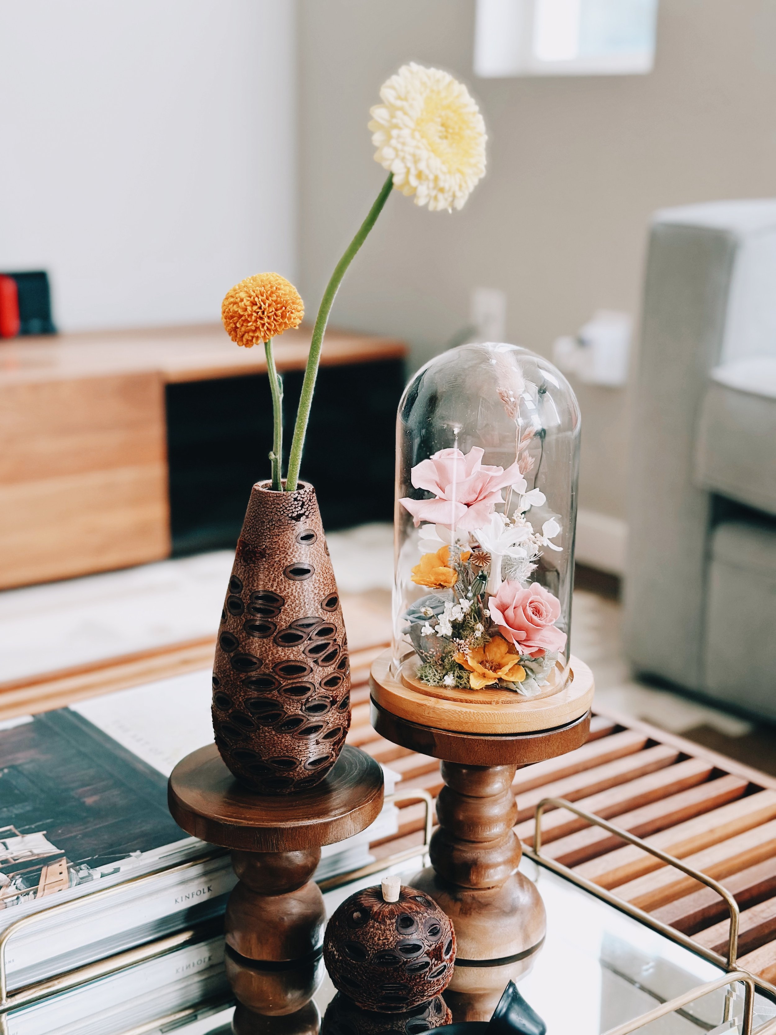 Decorative arrangement of artificial flowers in a clear glass dome and a tall textured vase on small wooden stands in a modern living room.
