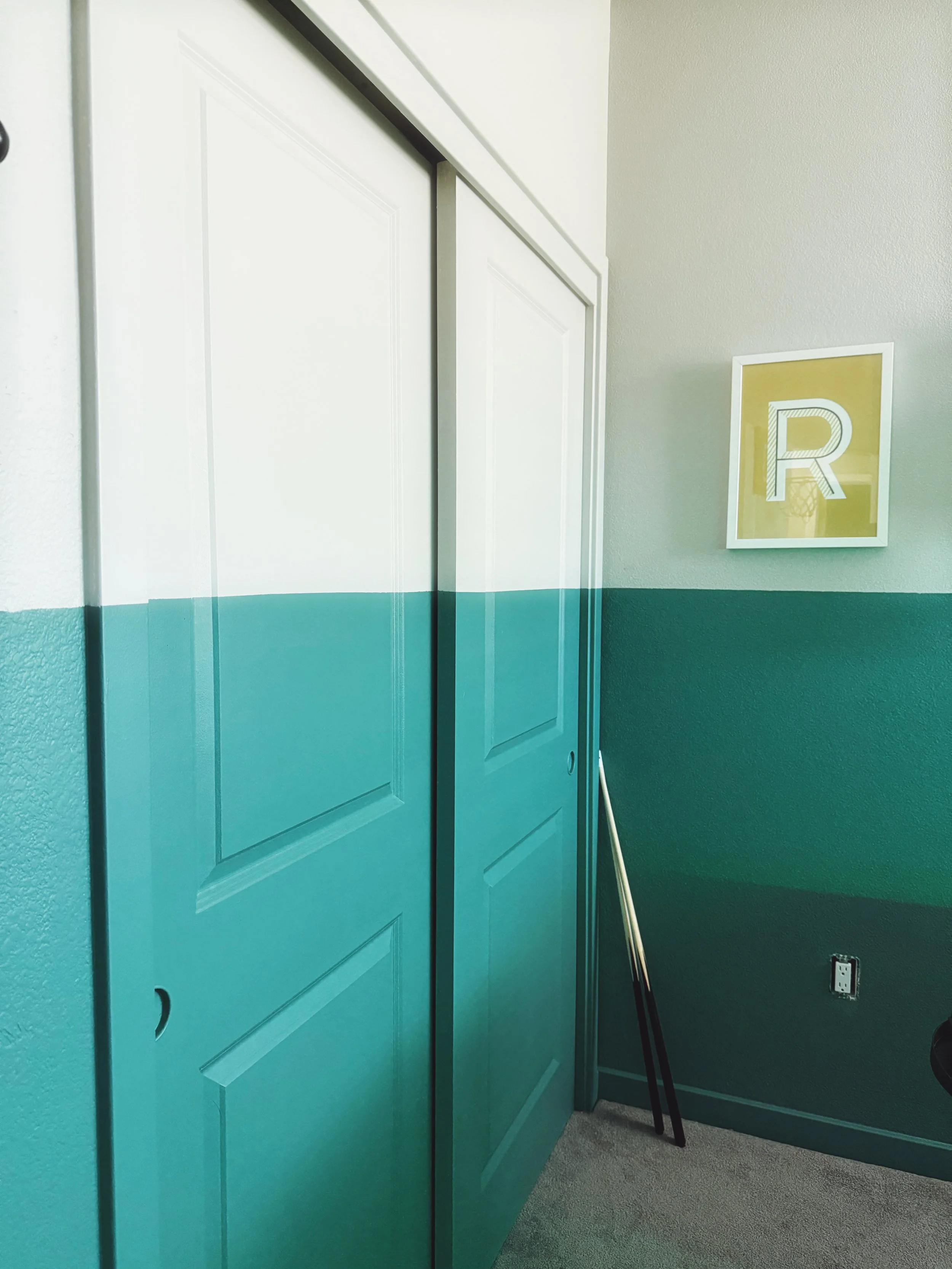 View of a partially open teal-colored closet door, broom with a black handle leaning against the wall, framed letter 'R' artwork on the wall, electrical outlet on the wall, and beige carpeting on the floor.