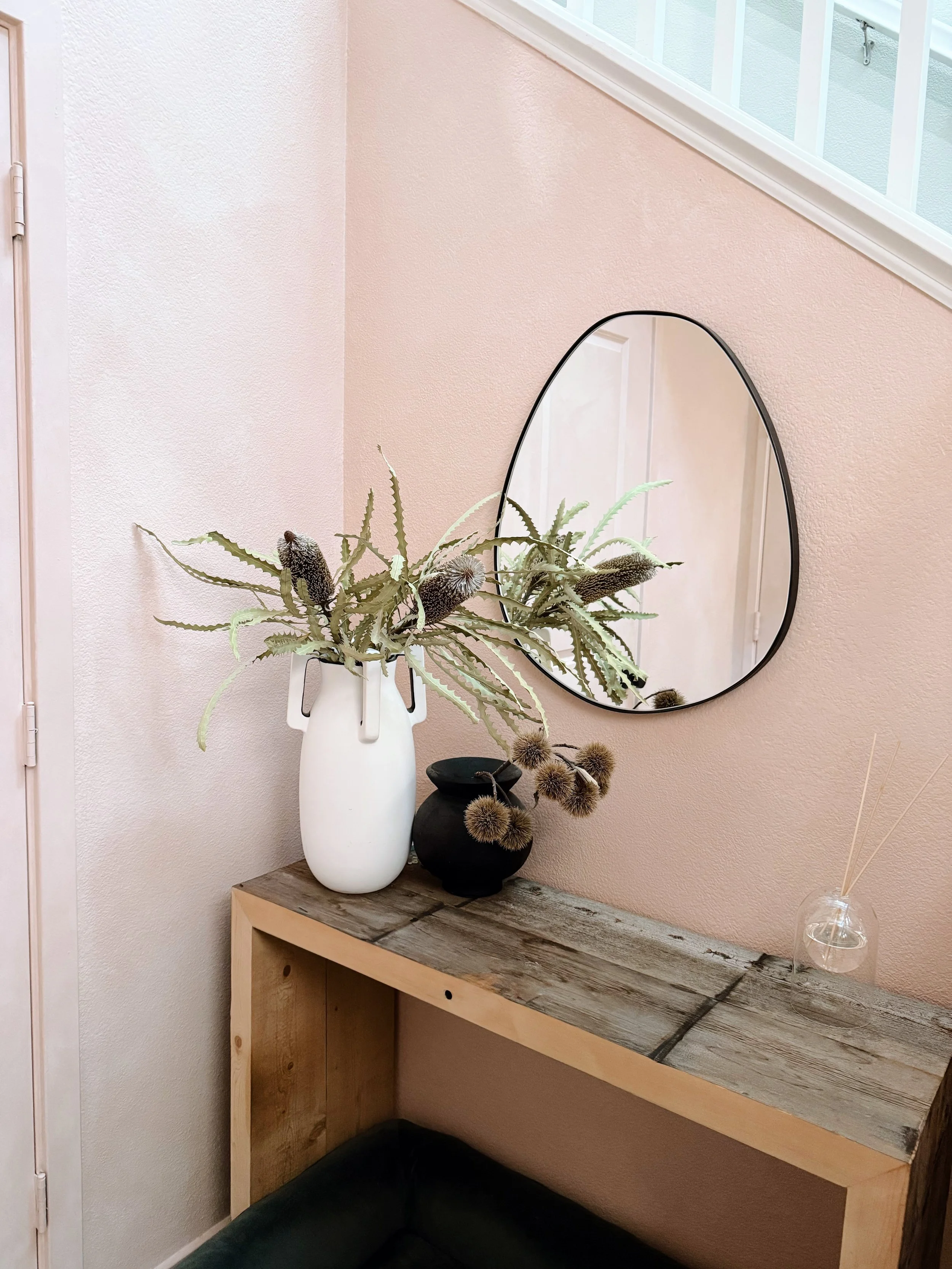 A corner of a room with a wooden table, a white vase with green and brown dried plants, a small black vase with dried thistles, a triangular mirror mounted on the wall, a glass diffuser with reed sticks, and a pink textured wall.