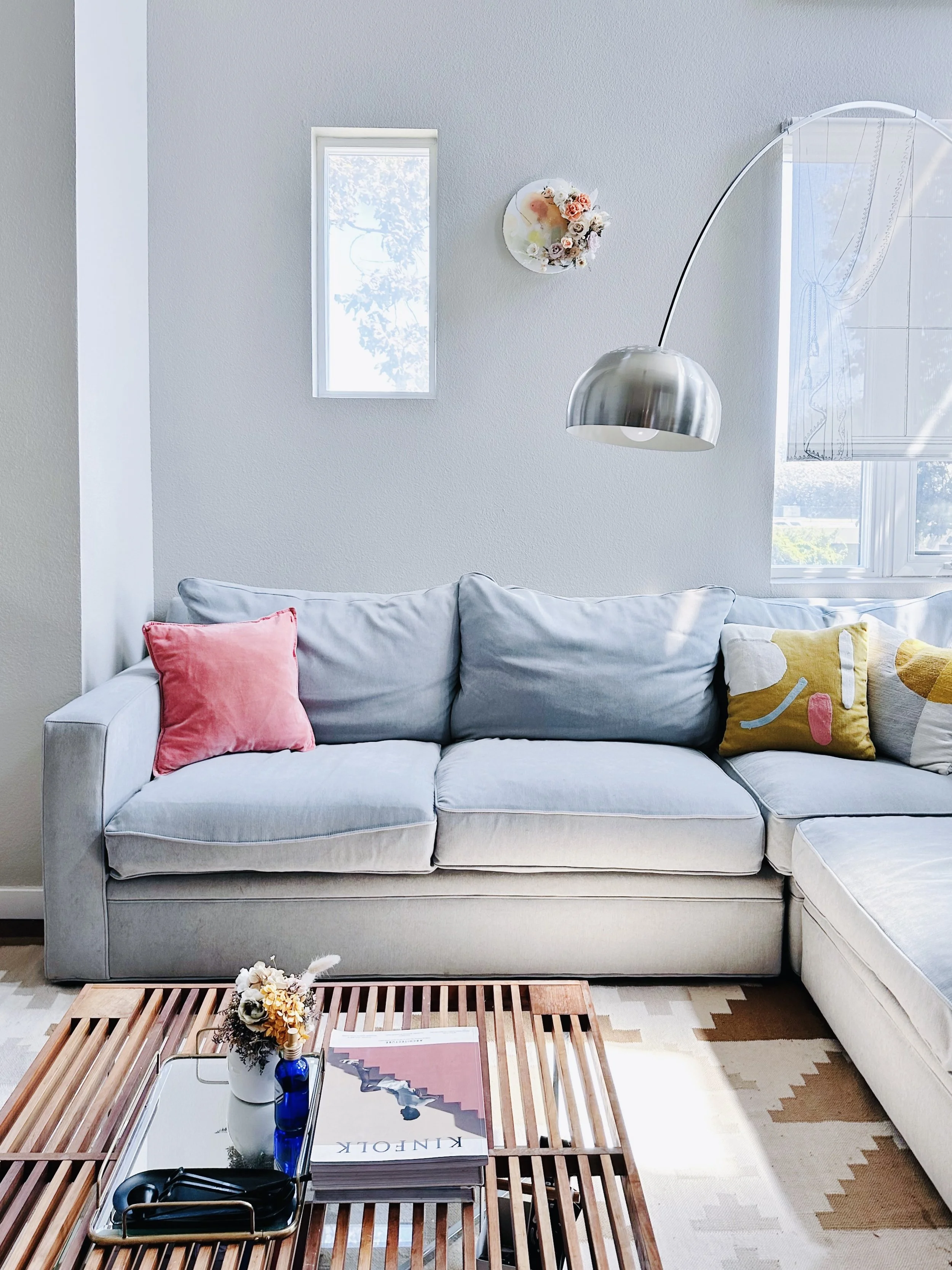 Living room with a light-colored sofa featuring pink, yellow, and patterned pillows, a coffee table with a magazine, decorative flowers, a tray with headphones, and a modern metallic desk lamp next to a window with sheer curtains. The wall has small windows and art decorations.