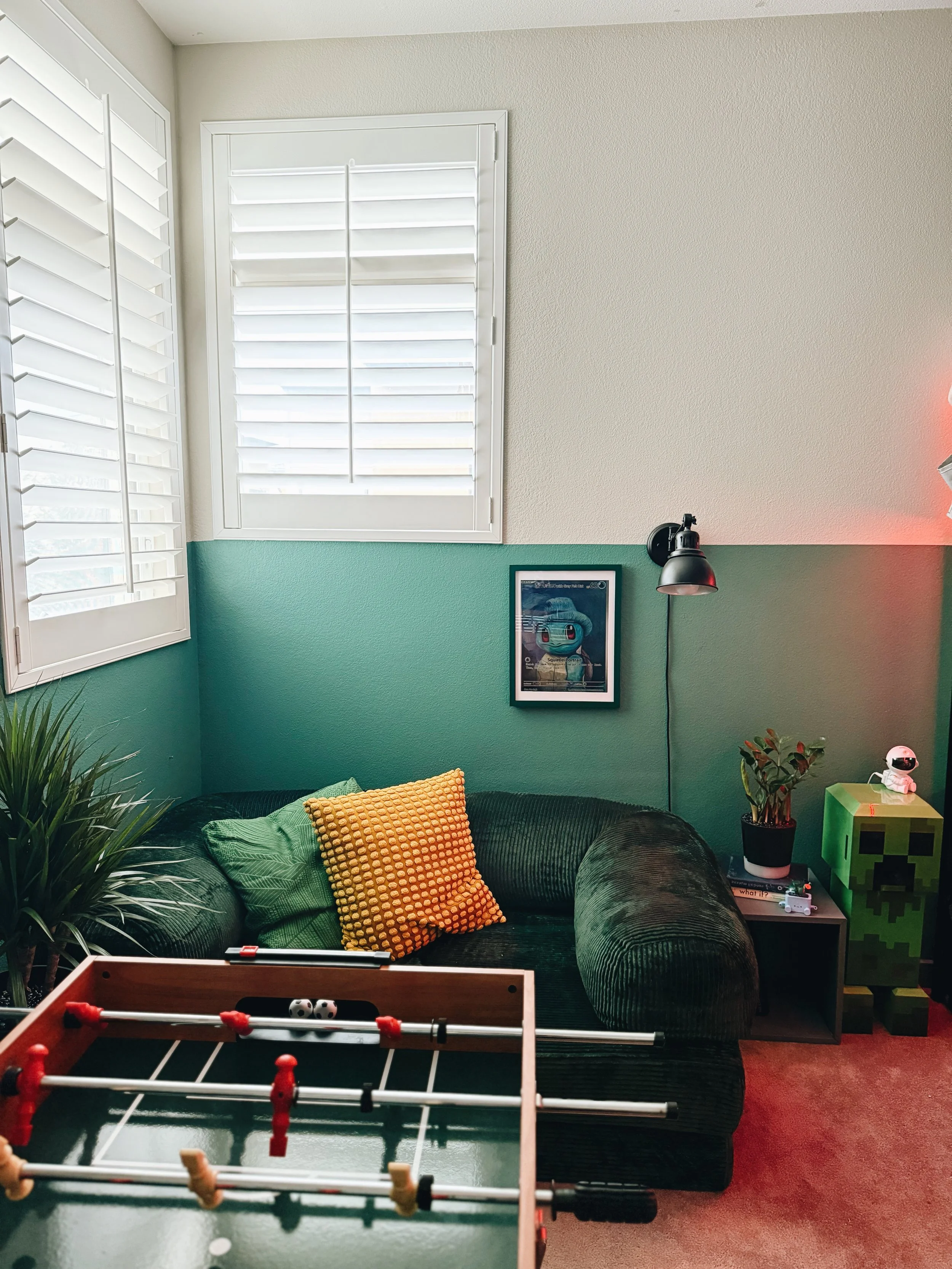 A cozy living room with a dark green velvet sofa, decorated with orange and green pillows, near two windows with white plantation shutters. There is a framed art piece on the wall, a black wall lamp, and a small side table with plants, a toy, and oth