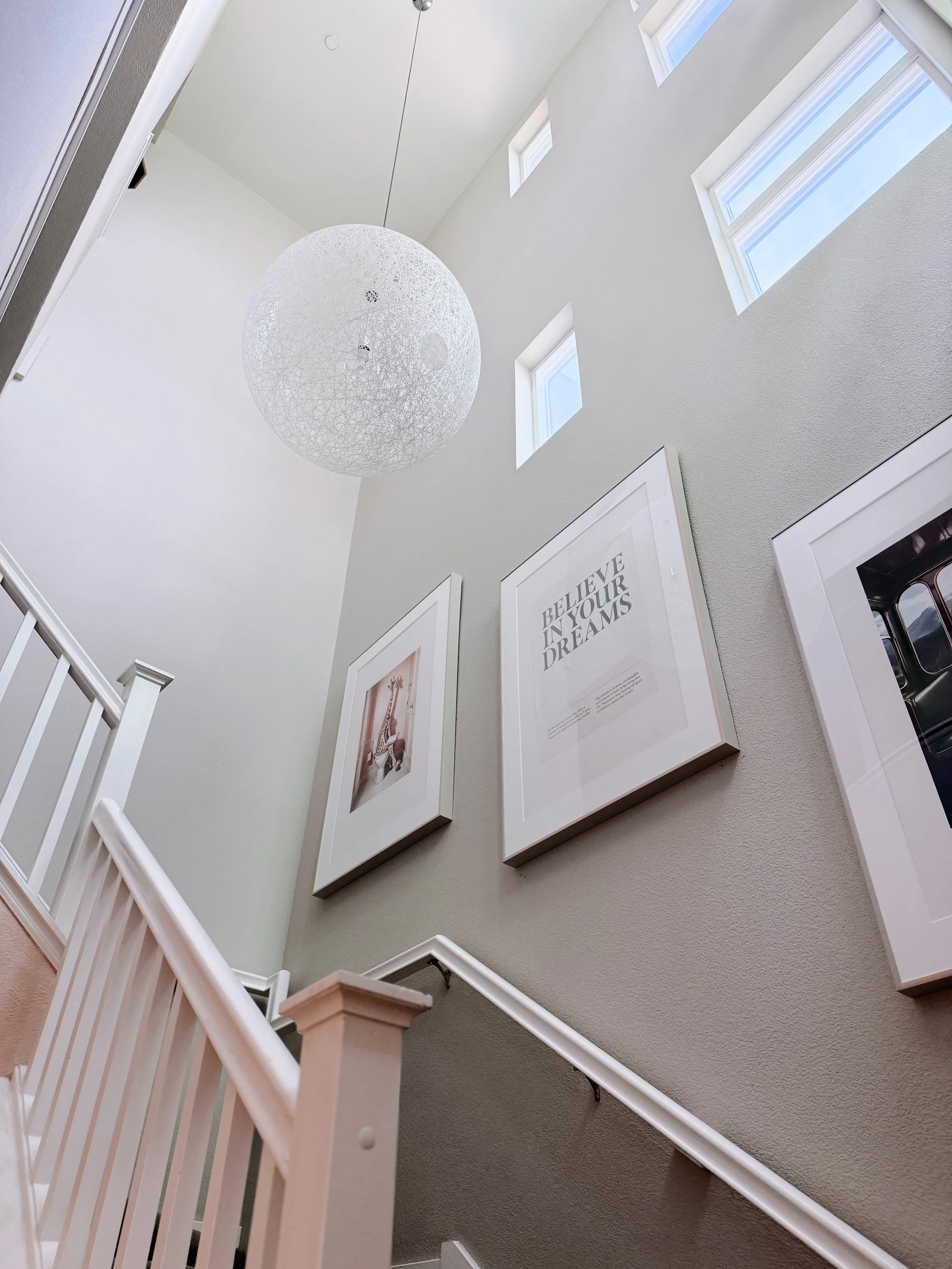 A modern staircase with a white railing leading up to a spacious, well-lit hallway with tall walls. Several framed artworks and motivational quotes are hung on the wall, and three small square windows let sunlight into the space. A distinctive, spher