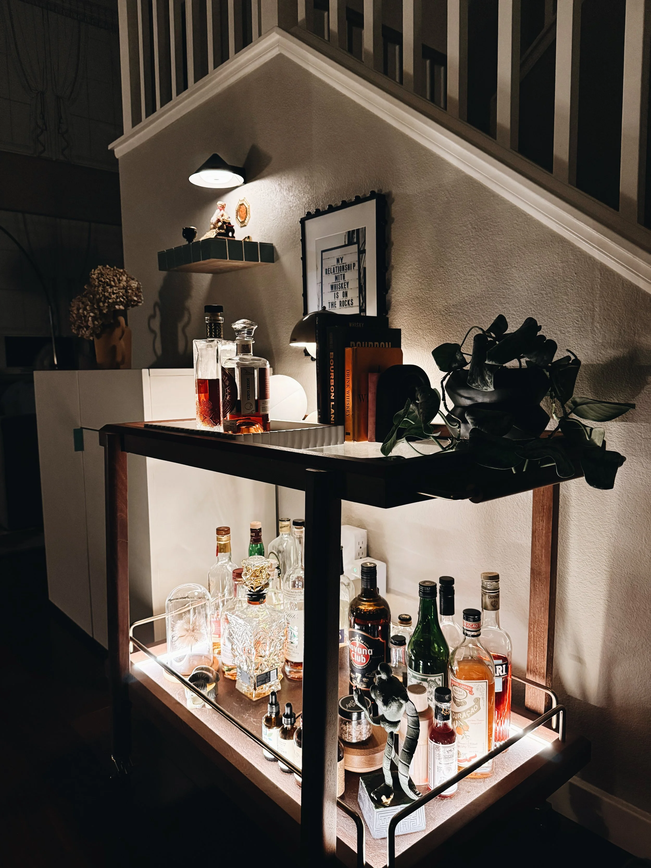 A lit bar cart with various bottles of alcohol, glasses, a small potted plant, and decorative items against a wall with books and a small shelf, under a wall-mounted light.