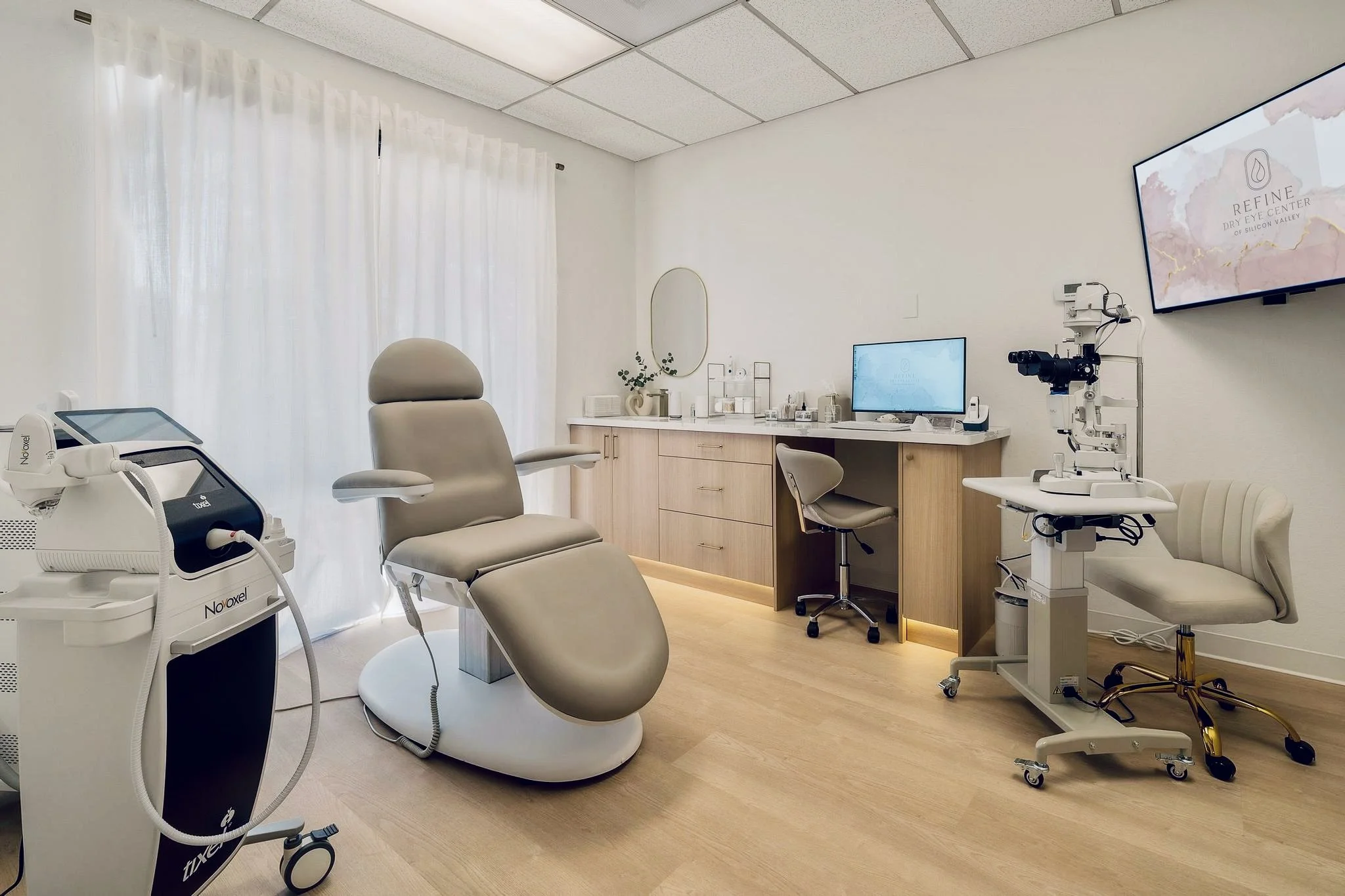 A medical examination room with a reclining chair, medical equipment, a monitor on the wall, and a wooden cabinet with supplies.