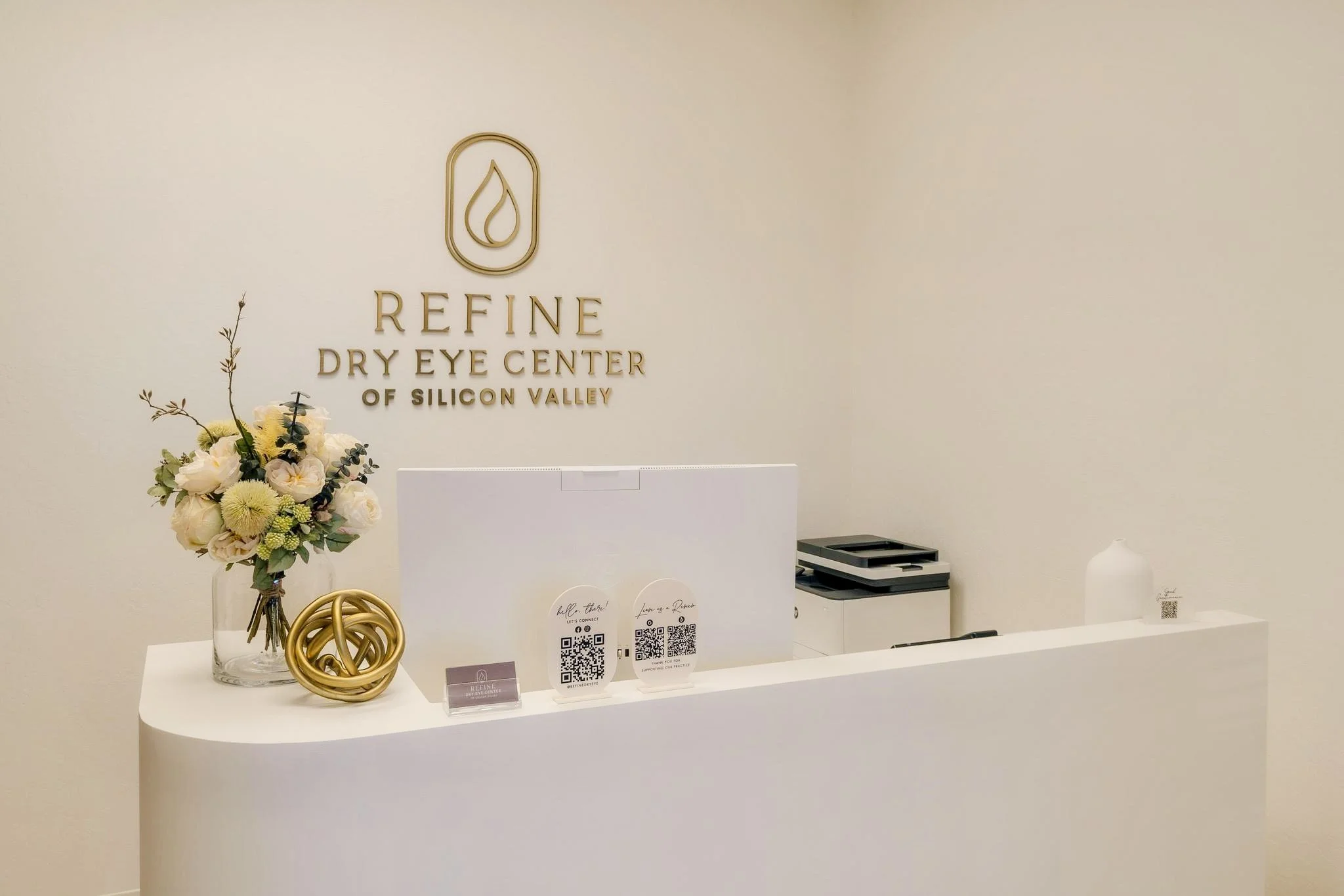 Reception desk at Reine Dry Eye Center of Silicon Valley with a floral arrangement, decorative gold object, QR code signs, and a white wall with the clinic's logo and name.