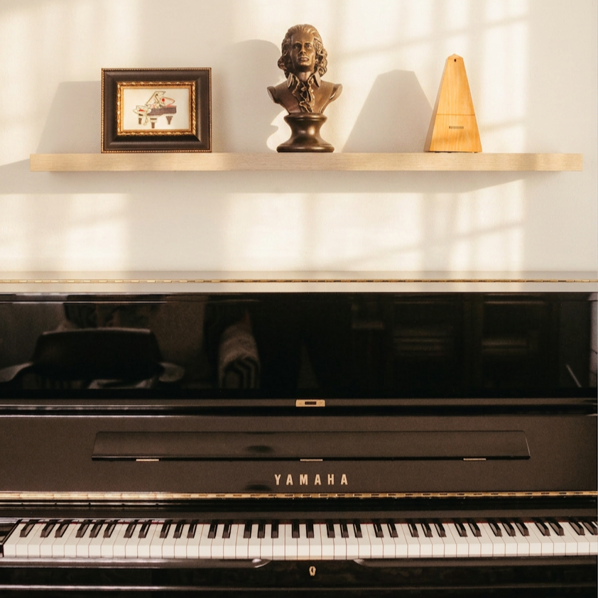 A black Yamaha piano with a closed lid, placed against a wall with a floating wooden shelf above it. On the shelf, from left to right, there is a framed picture of a grand piano, a bust sculpture of a composer, and a wooden metronome.