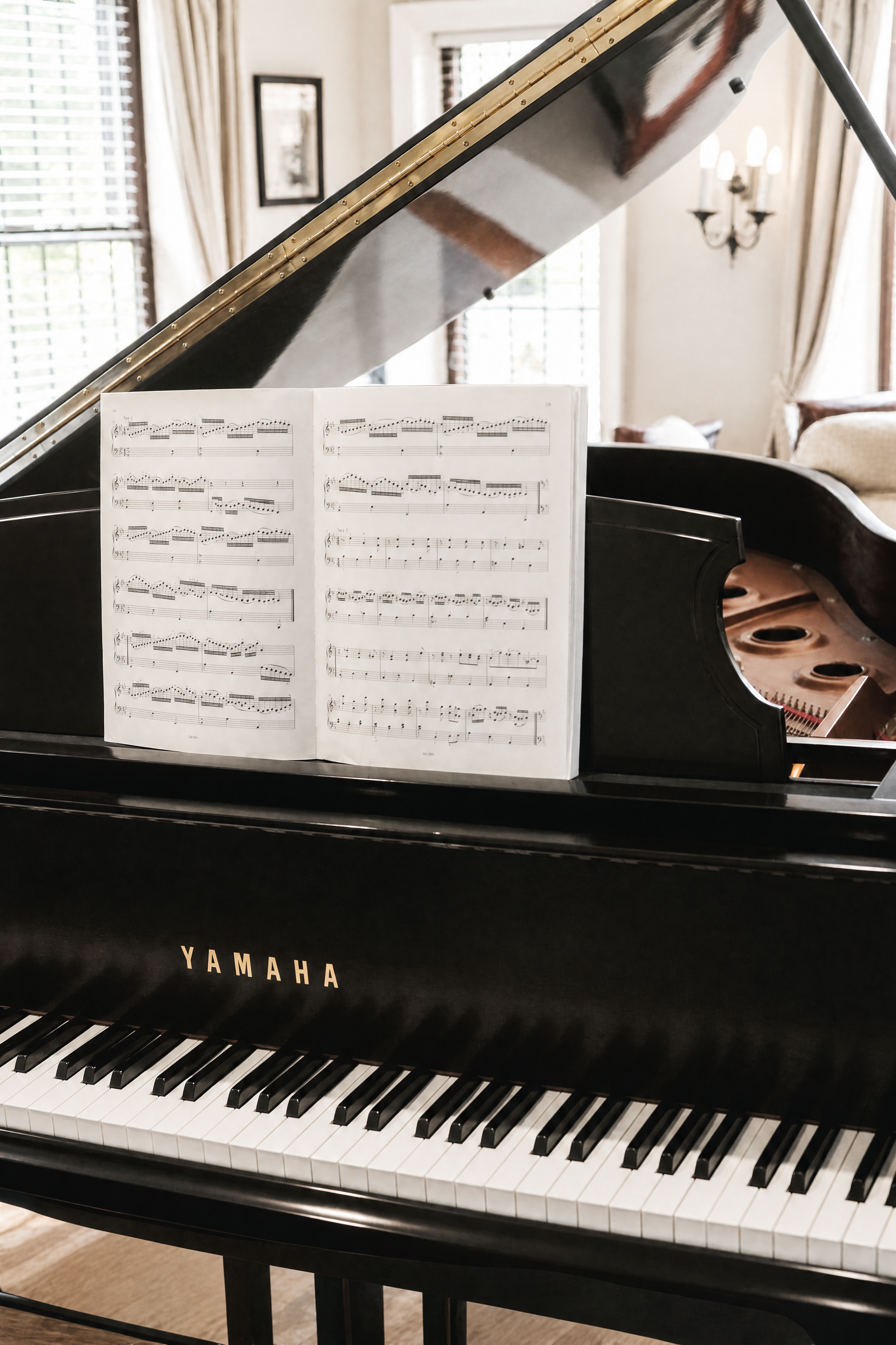 A black Yamaha grand piano with an open sheet music stand showing sheet music, in a well-lit room with windows, curtains, and a chandelier.