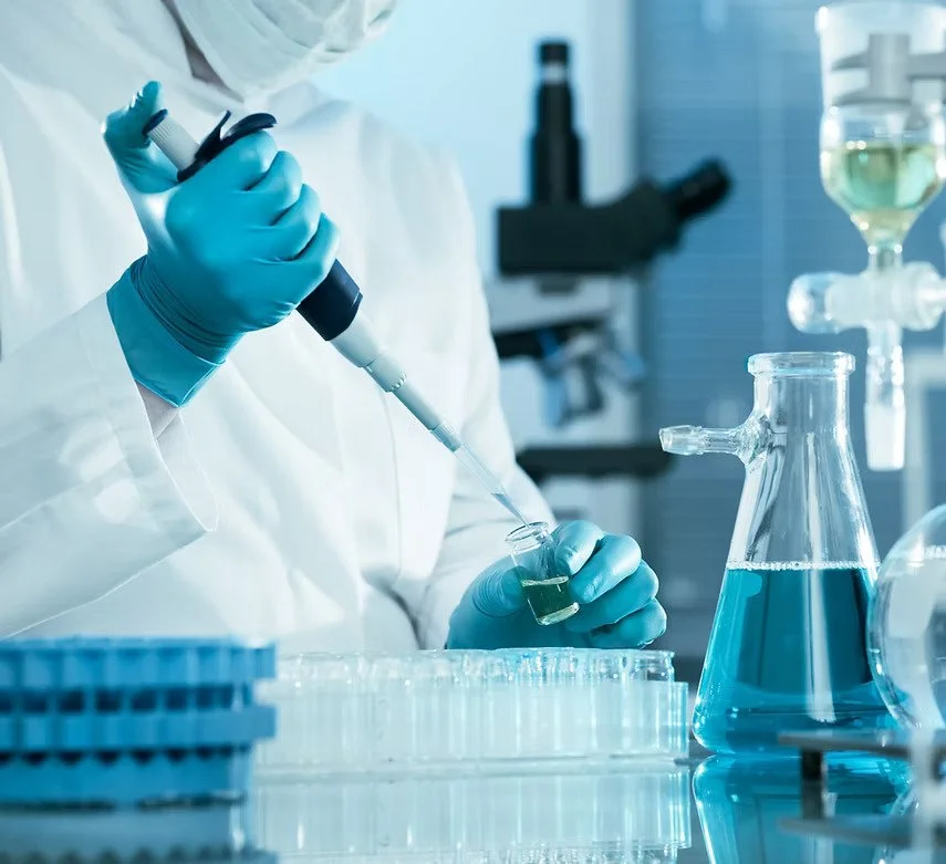 Scientist wearing gloves using a pipette to transfer liquid into a test tube in a laboratory setting with glassware and scientific equipment.