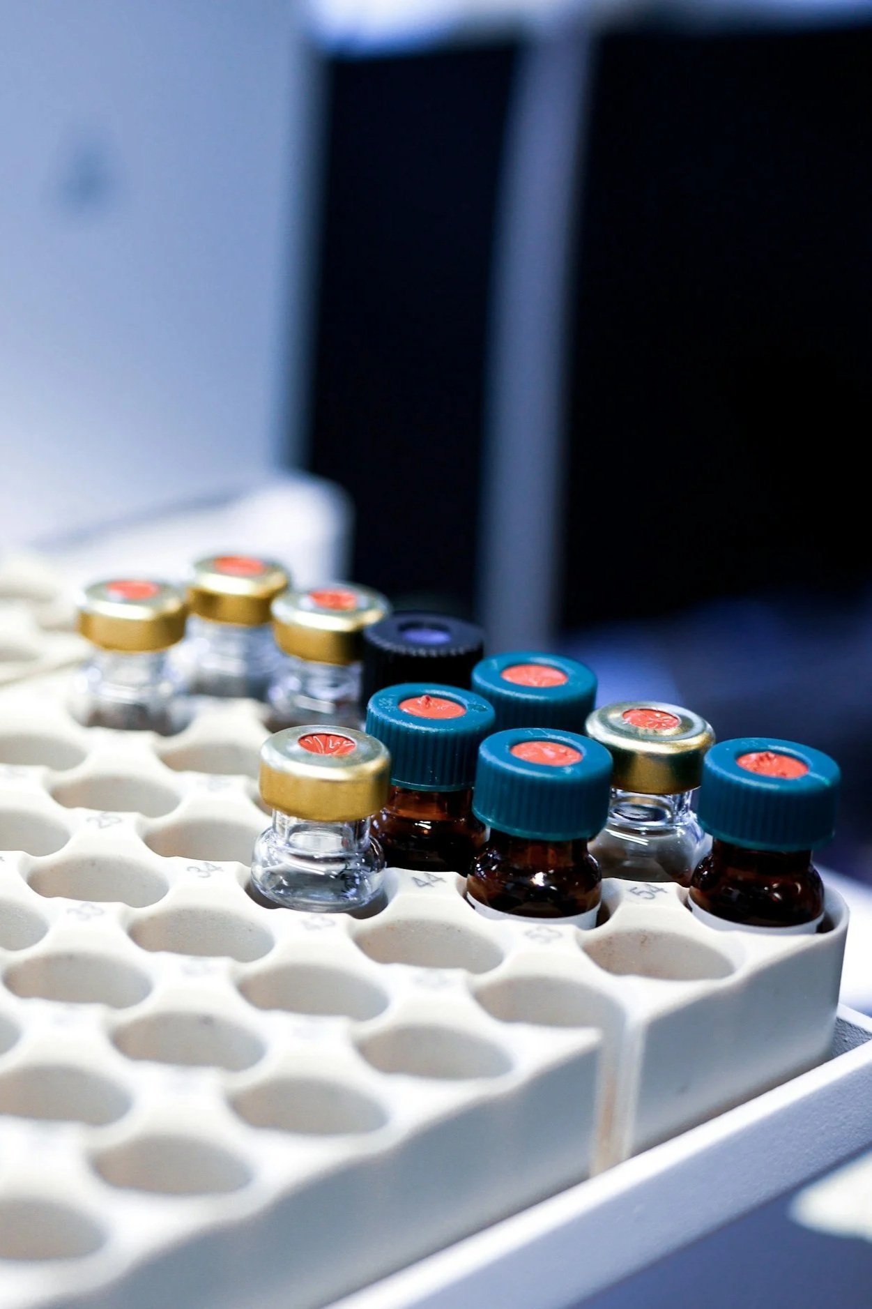 Close-up of glass vials with colored caps in a white foam tray, in a laboratory setting.