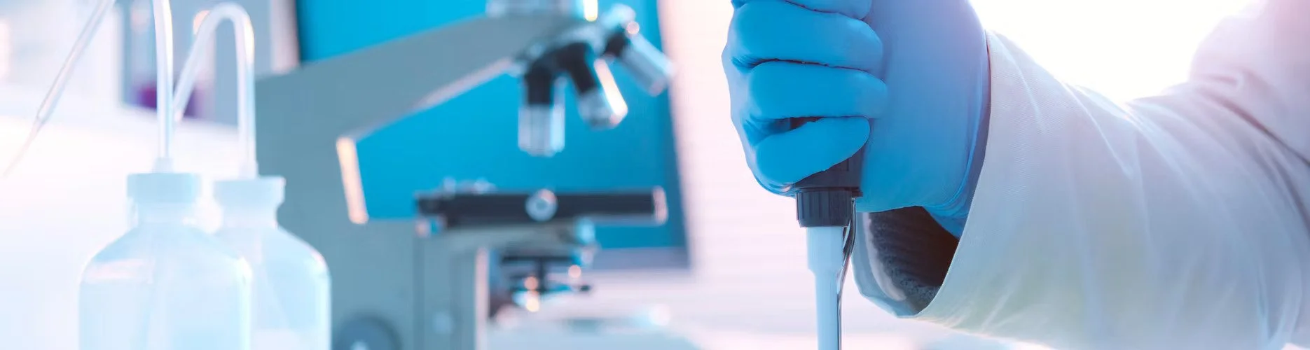 A scientist in a lab using a micropipette to transfer liquid into a test tube, with laboratory equipment and bottles visible in the background.