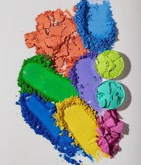 A colorful collection of powdered pigments in various shades including orange, blue, green, purple, yellow, and pink, with some larger spherical pigment chunks.
