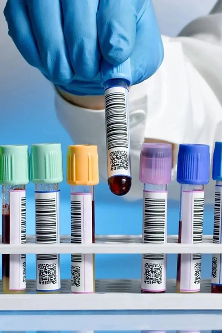 A healthcare professional wearing blue gloves handles a blood sample tube with a barcode, among other blood sample tubes with colored caps in a rack.