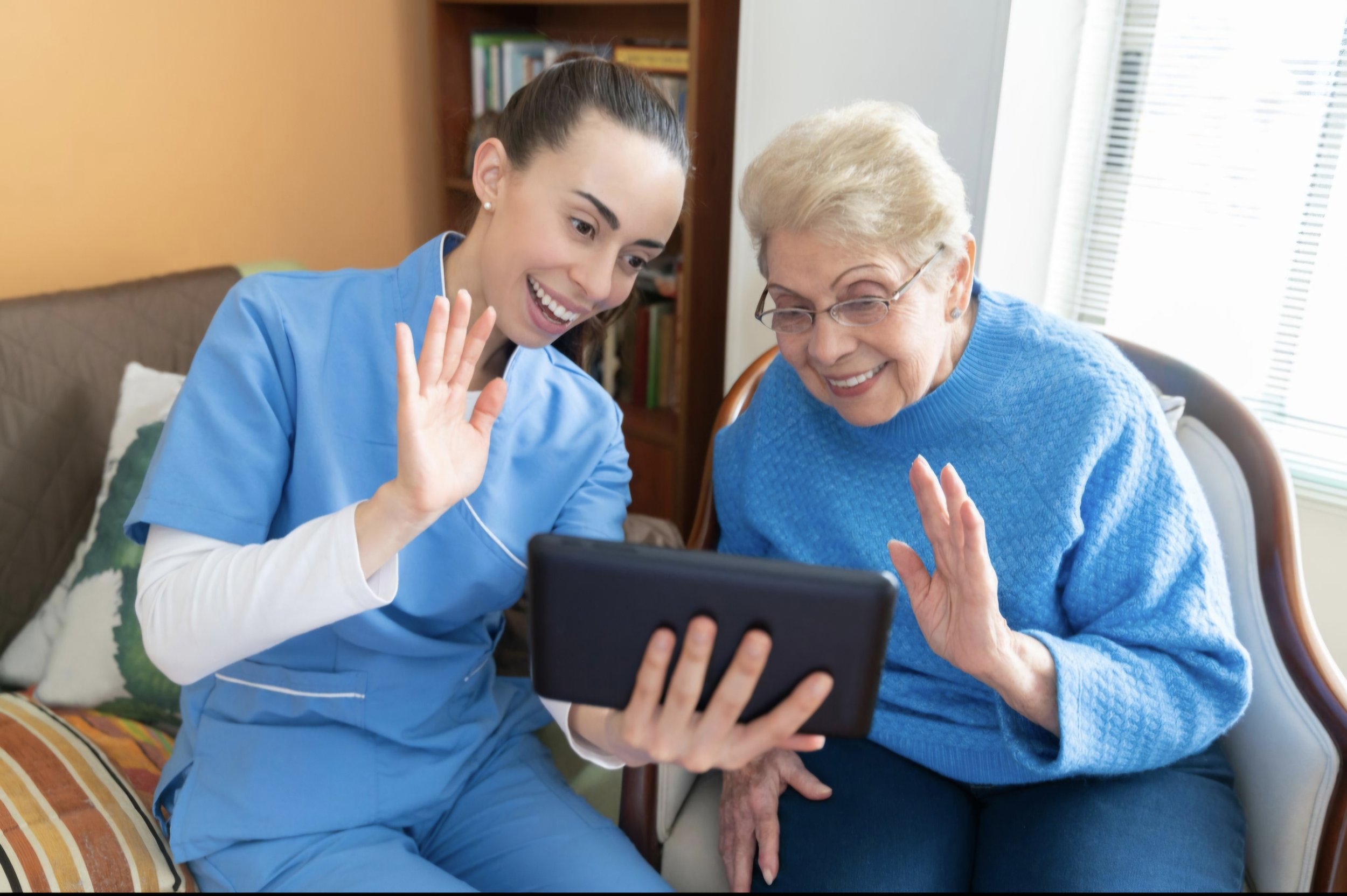 A young woman in medical scrubs and an elderly woman are sitting together, smiling and waving at a tablet, likely video chatting, in a cozy room with a window and a bookshelf in the background.
