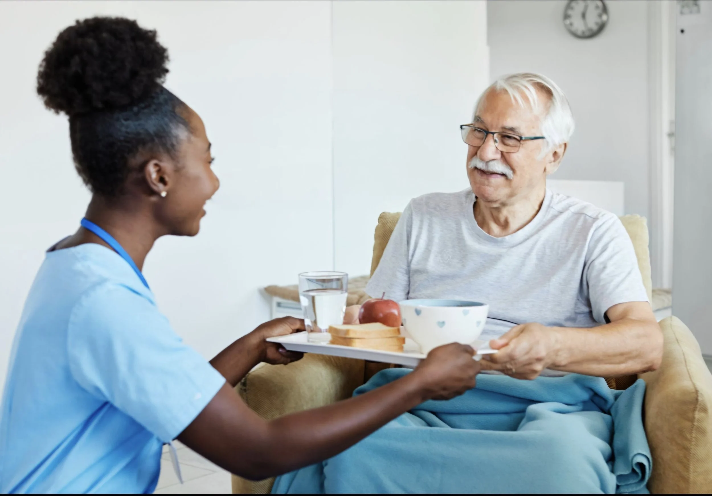 A healthcare worker in blue scrubs holding a tray with a glass of water, slices of bread, and an apple, serving an elderly man with white hair and glasses, who is smiling and sitting on a yellow chair during a meal in a bright room with a clock on the wall.