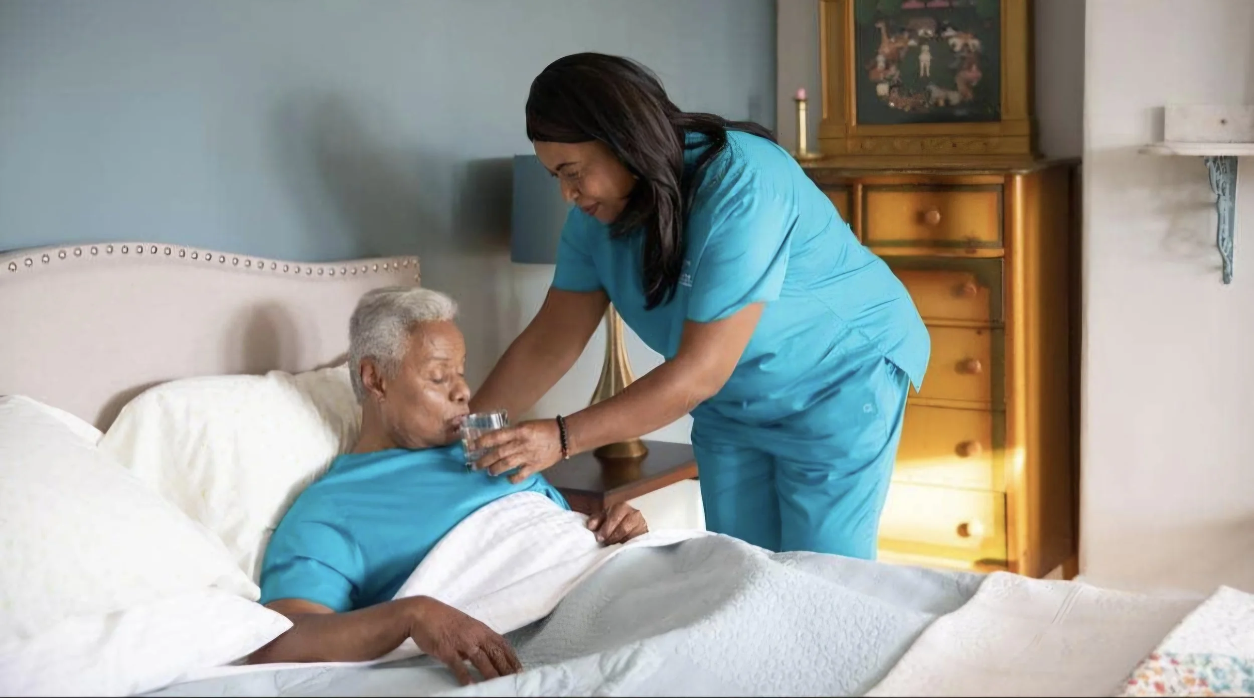 A healthcare worker in blue scrubs offers a glass of water to an elderly patient lying in bed, smiling with his eyes closed.