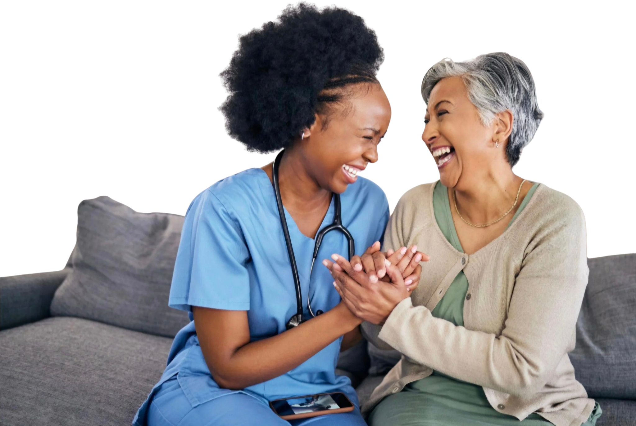 A nurse and an elderly woman sharing a joyful moment on a couch, holding hands, with the nurse wearing blue scrubs and a stethoscope around her neck.