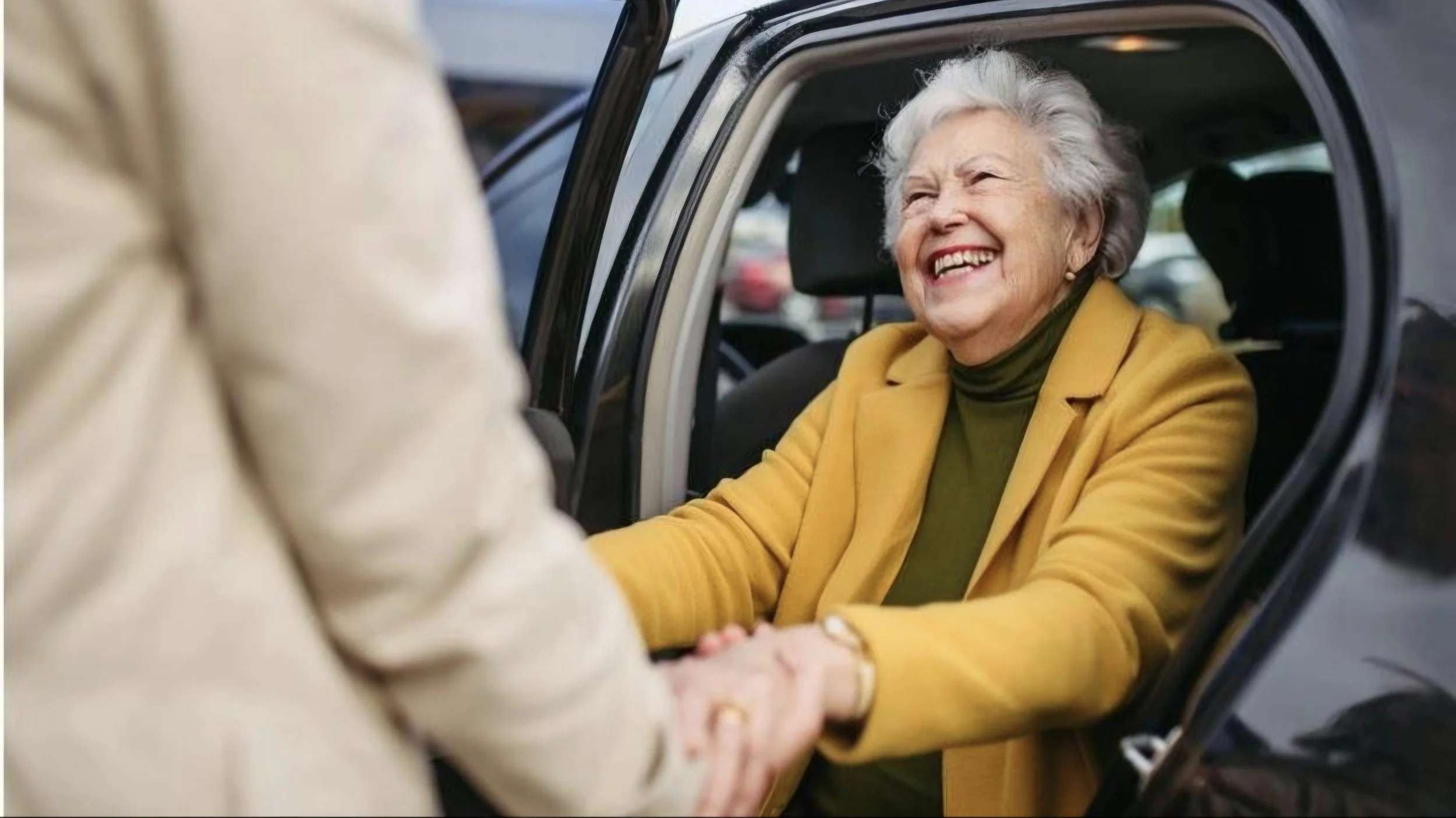 An elderly woman with gray hair, wearing a mustard yellow coat, smiling warmly while sitting in a car and holding hands with a person outside the car.