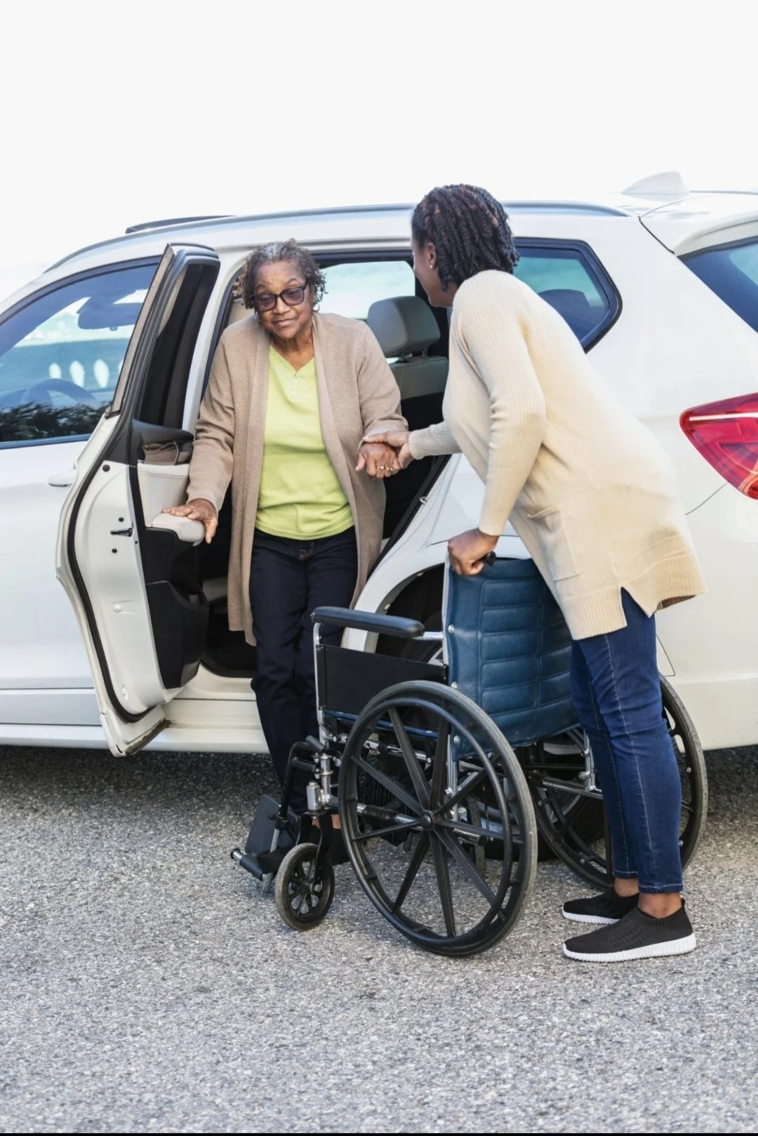 A woman is helping another woman, who is in a wheelchair, exit a white SUV. The woman in the wheelchair is wearing a beige cardigan and glasses, while the other woman is in a cream sweater, holding her hand, and standing next to a blue wheelchair.