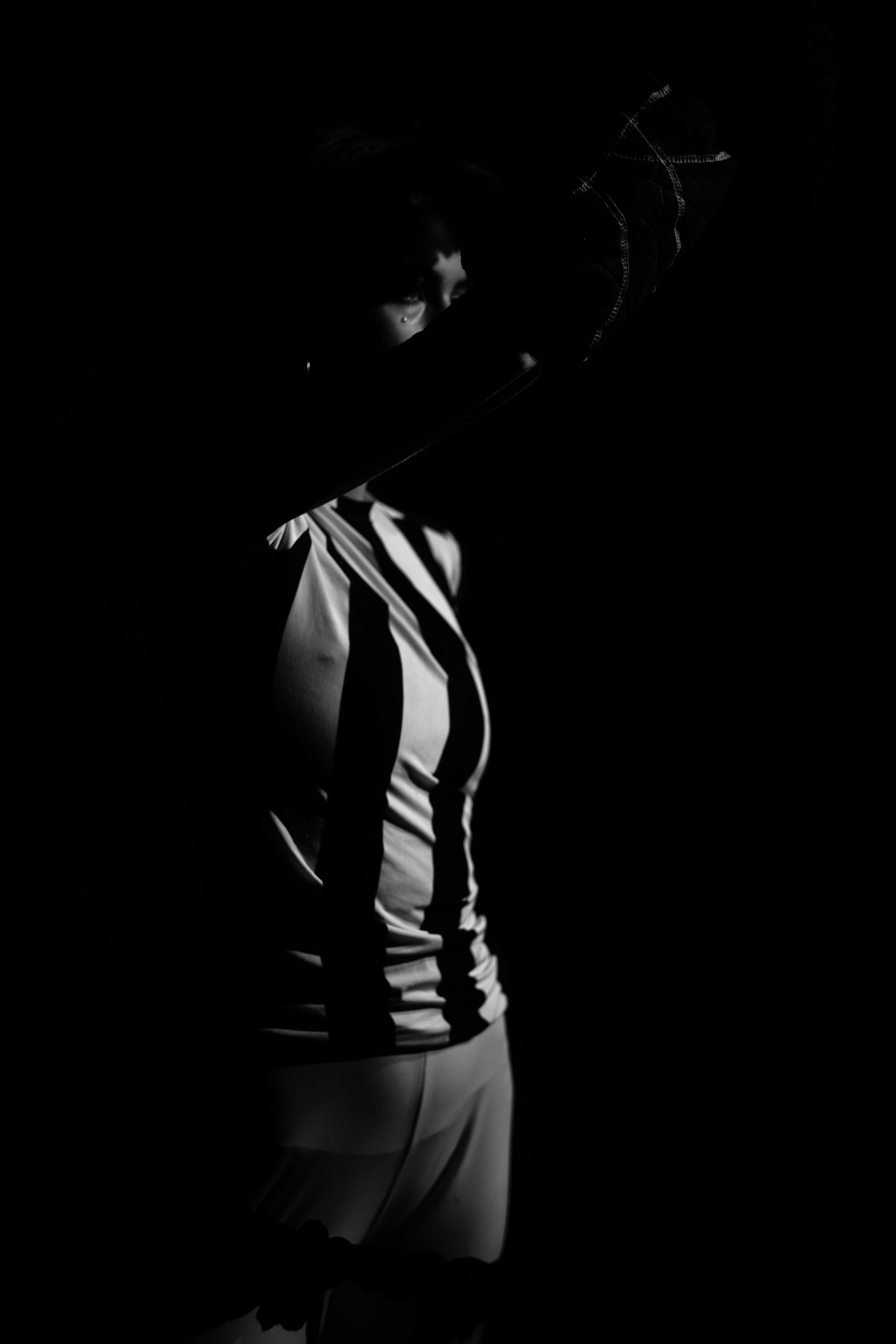 Eine Person in schwarz-weiß gestreiftem Top und weißen Hose vor schwarzem Hintergrund, teilweise im Schatten, mit Blick nach oben.