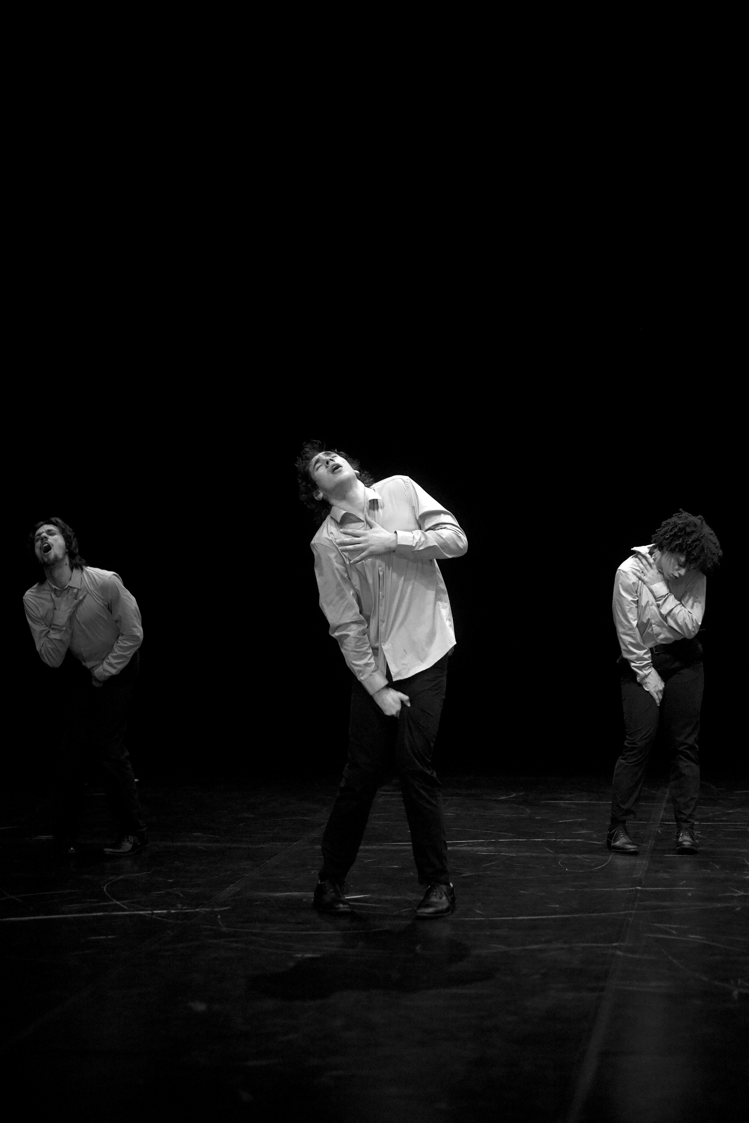 Drei männliche Tänzer führen eine moderne Tanzperformance auf einer Bühne vor einem schwarzen Hintergrund auf, alle tragen helle Hemden und dunkle Hosen.