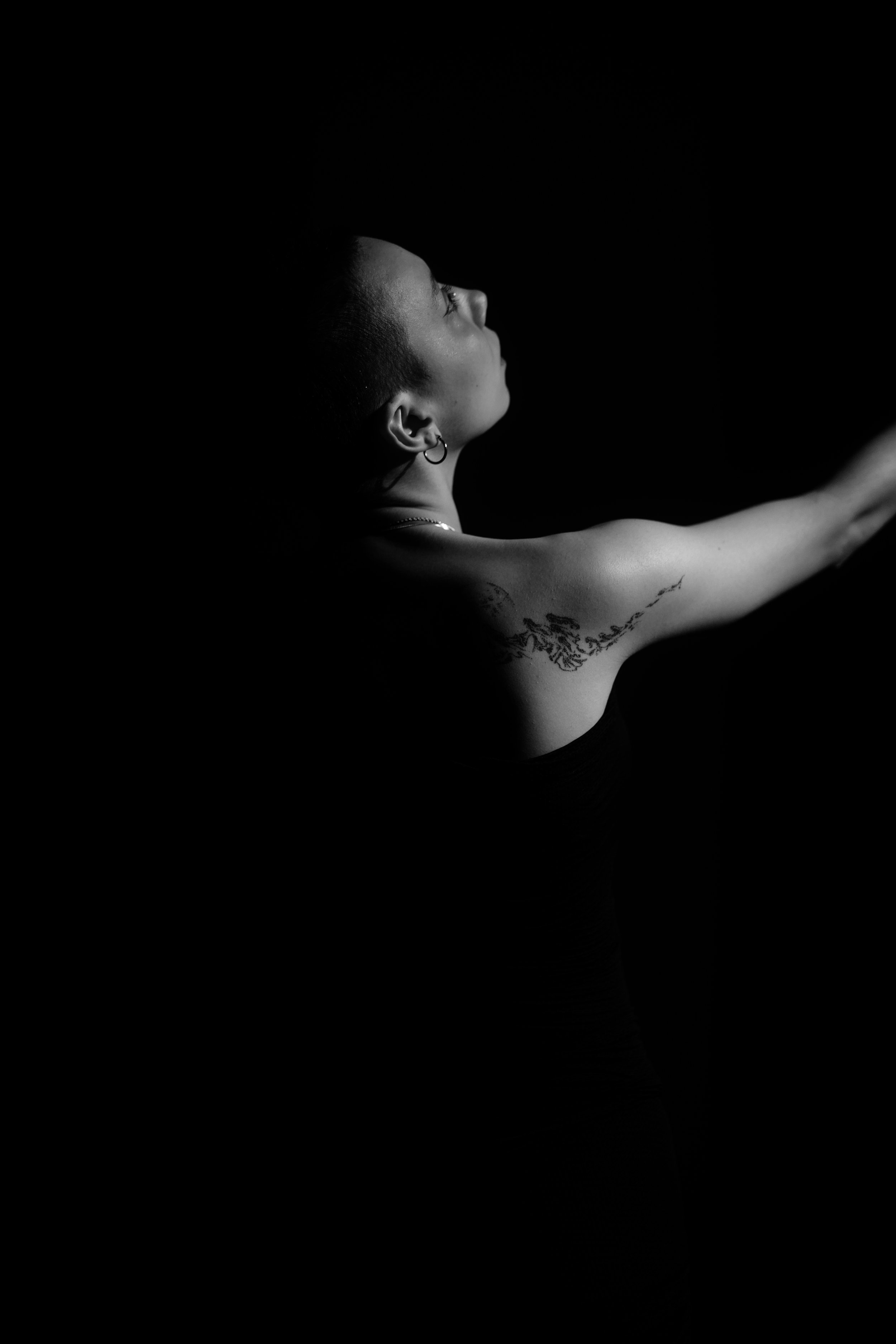 Schwarz-weiß-Foto eines Menschen, seitlich betrachtet, mit kurzem Haar, Tätowierungen am Arm und Piercing im Ohr. Der Arm ist ausgestreckt, der Hintergrund ist dunkel.