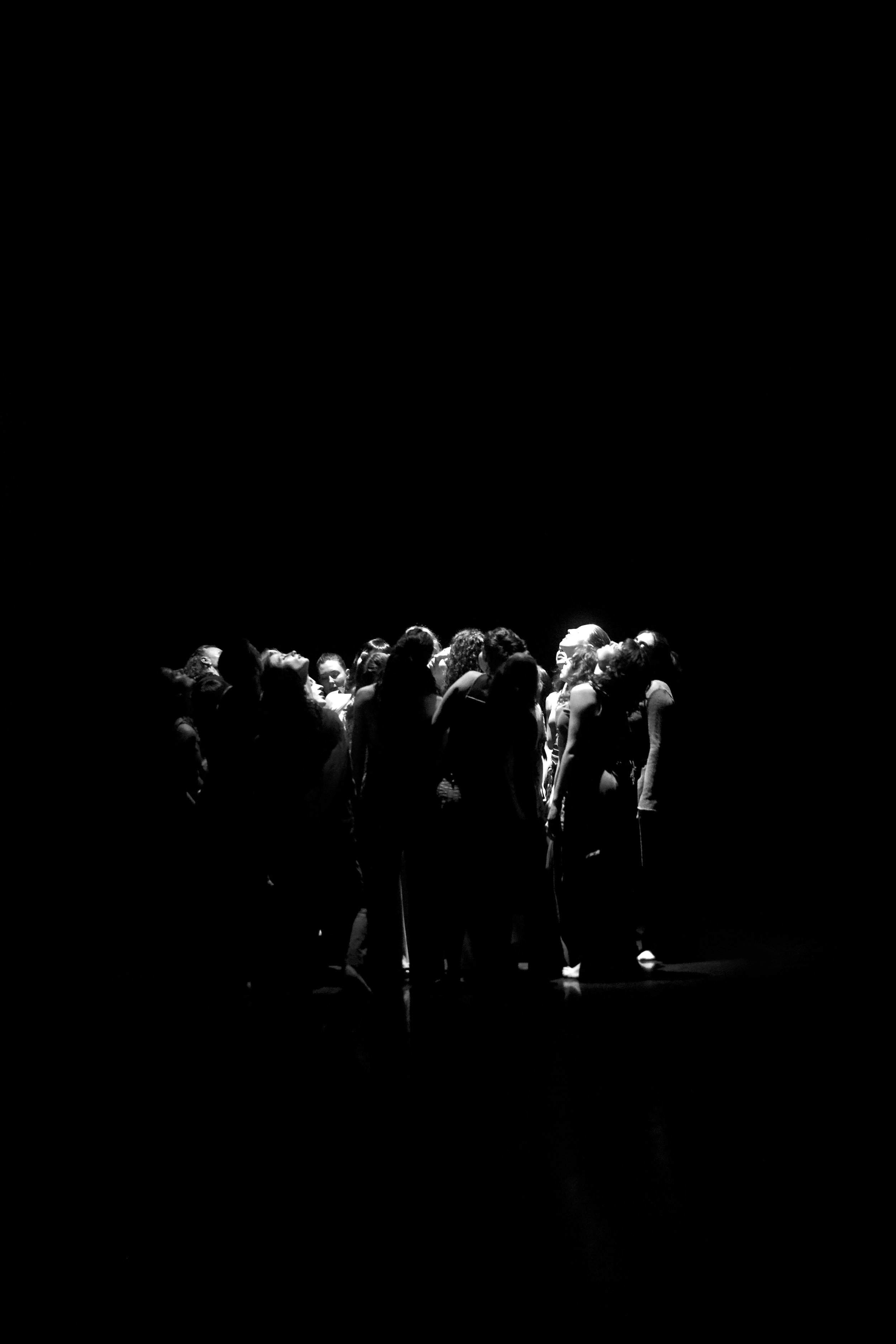 Eine Menschenmenge steht eng beieinander auf einer Bühne, nur ihre Köpfe sind sichtbar. Das Bild ist schwarz-weiß und die Szene scheint in einem Theater oder bei einer Veranstaltung stattgefunden zu haben.