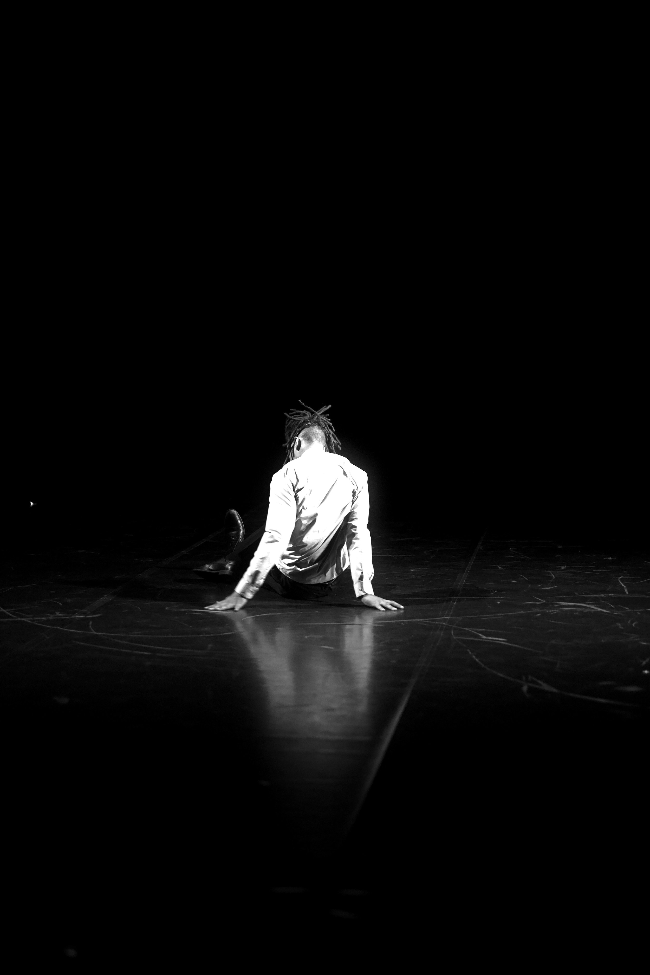 Ein Mensch sitzt auf der Bühne mit gestreiftem weißen Hemd und dunklen Schuhen, scheinbar in einer Tanz- oder Theaterperformance, die Situation ist dramatisch gestaltet, dunkler Hintergrund, Spotlight auf die Person