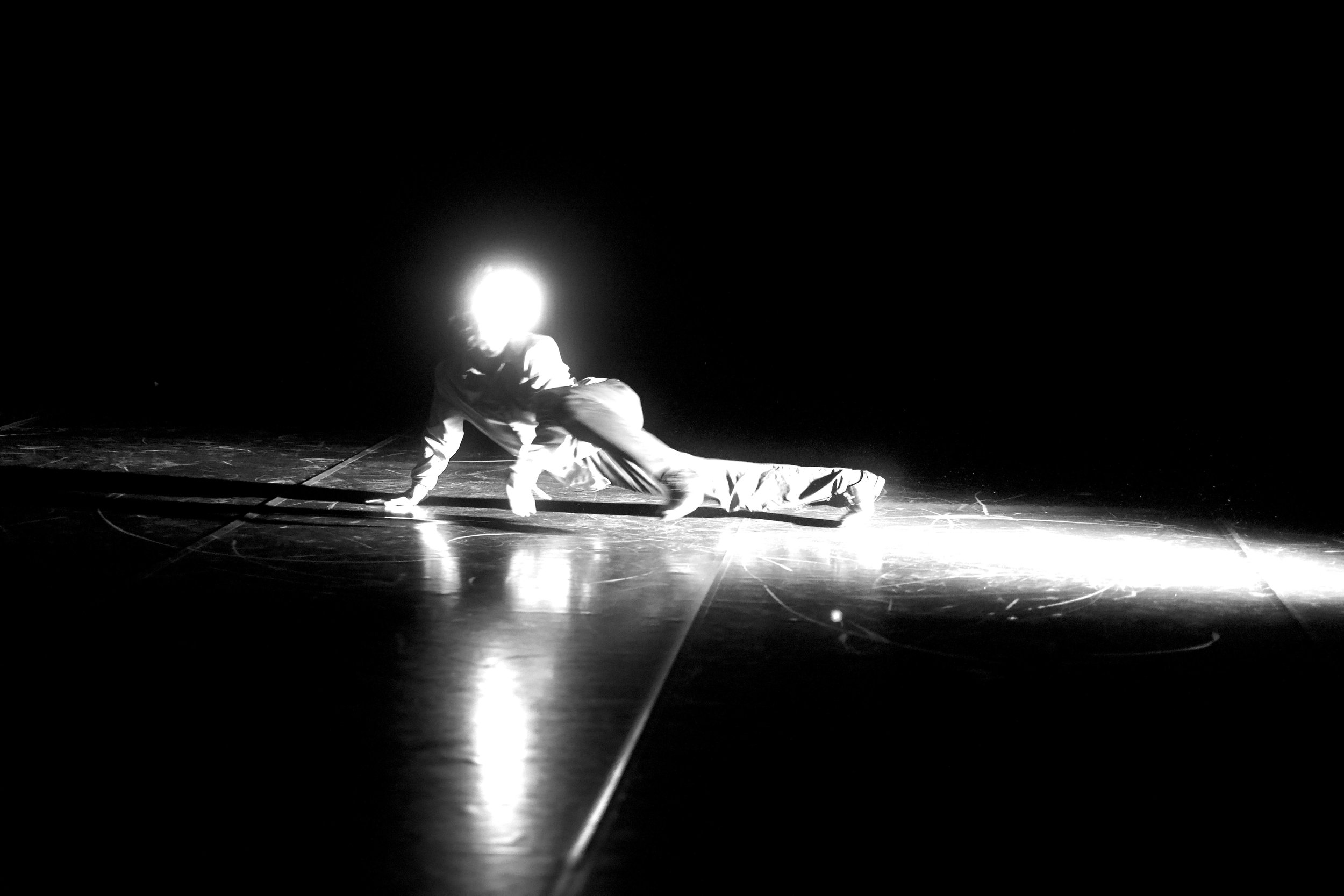 Eine Person auf einem dunklen Bühnenboden, die sich in einer Pose bewegt, beleuchtet von hellem Licht aus dem Hintergrund, wodurch die Silhouette hervorgehoben wird.