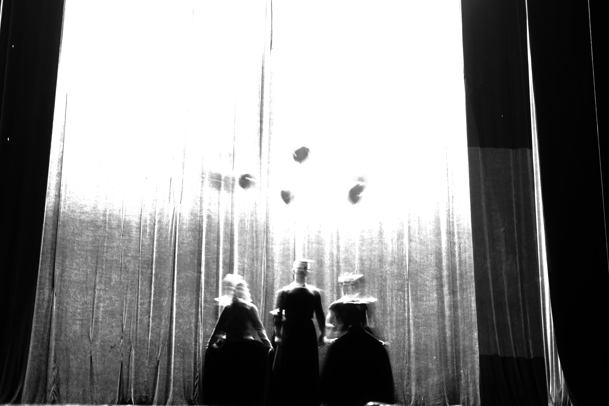 Drei Kinder sitzen auf einer Bühne oder im Theater, von hinten sichtbar, während sie einem roten Vorhang zuschauen, der an einer erhöhten Bühne hängt.