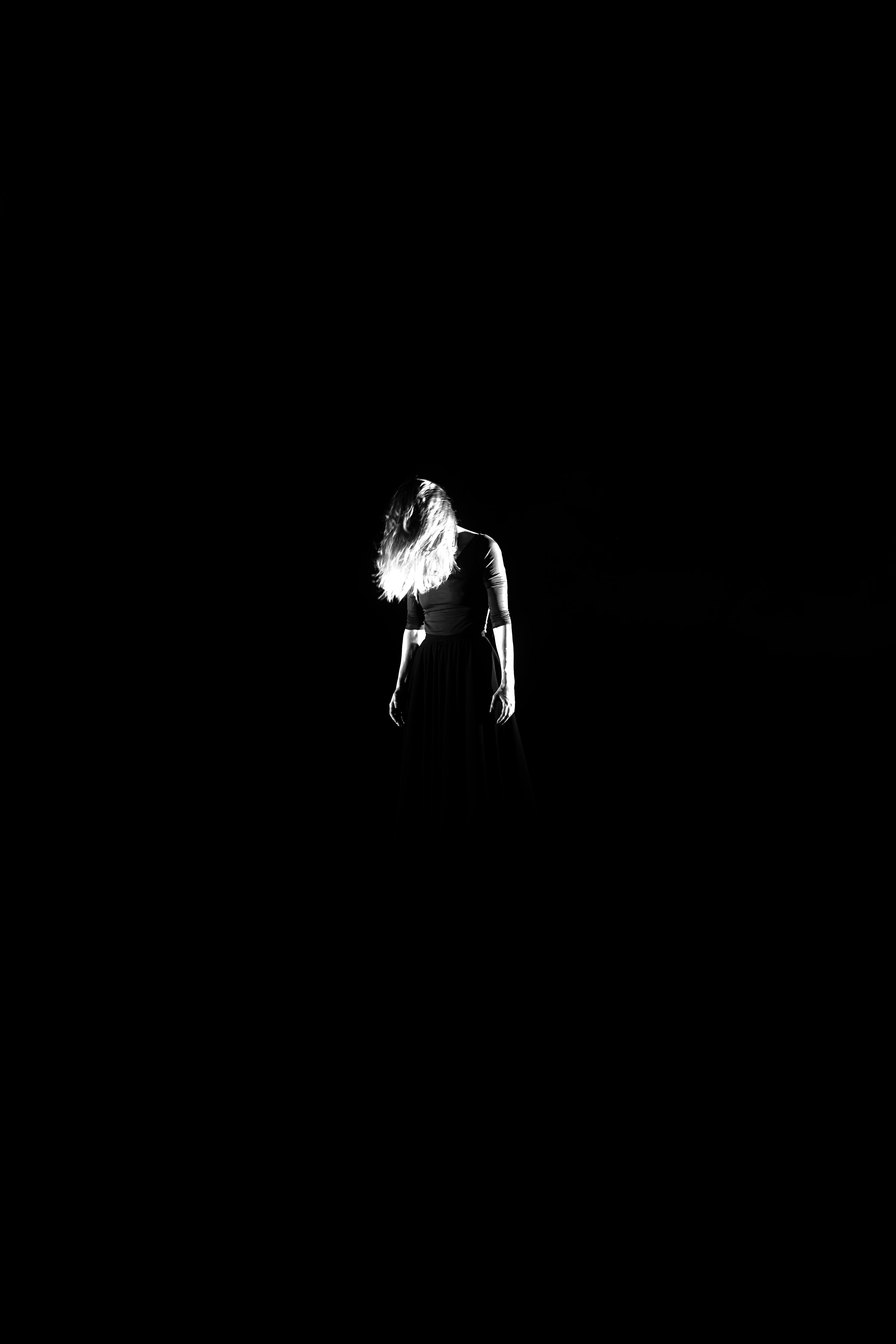 Schwarz-weiß-Foto eines Mädchens mit langen, lockigen Haaren, das in einem schwarzen Kleid steht, während es nach unten schaut, bei schwachem Licht, das einen dramatischen Effekt schafft.