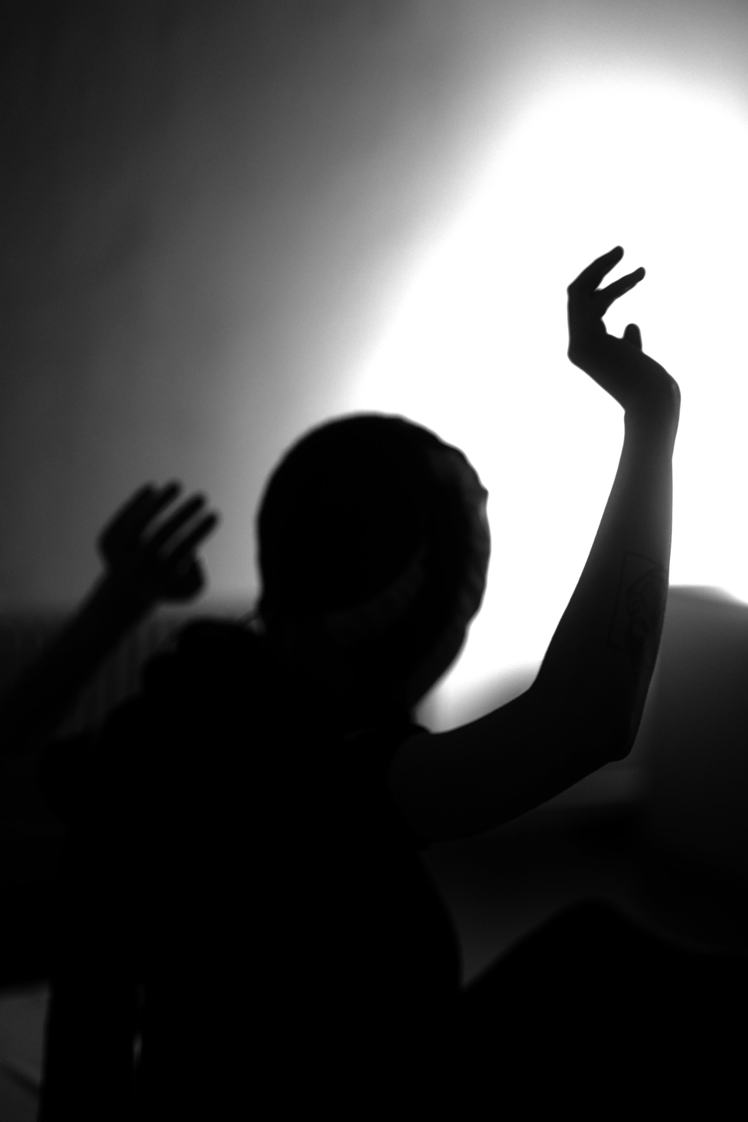 Schwarz-weißer Schatten eines Menschen mit erhobenen Händen vor einer hellen Lichtquelle, Kopf und Arme sind deutlich sichtbar.