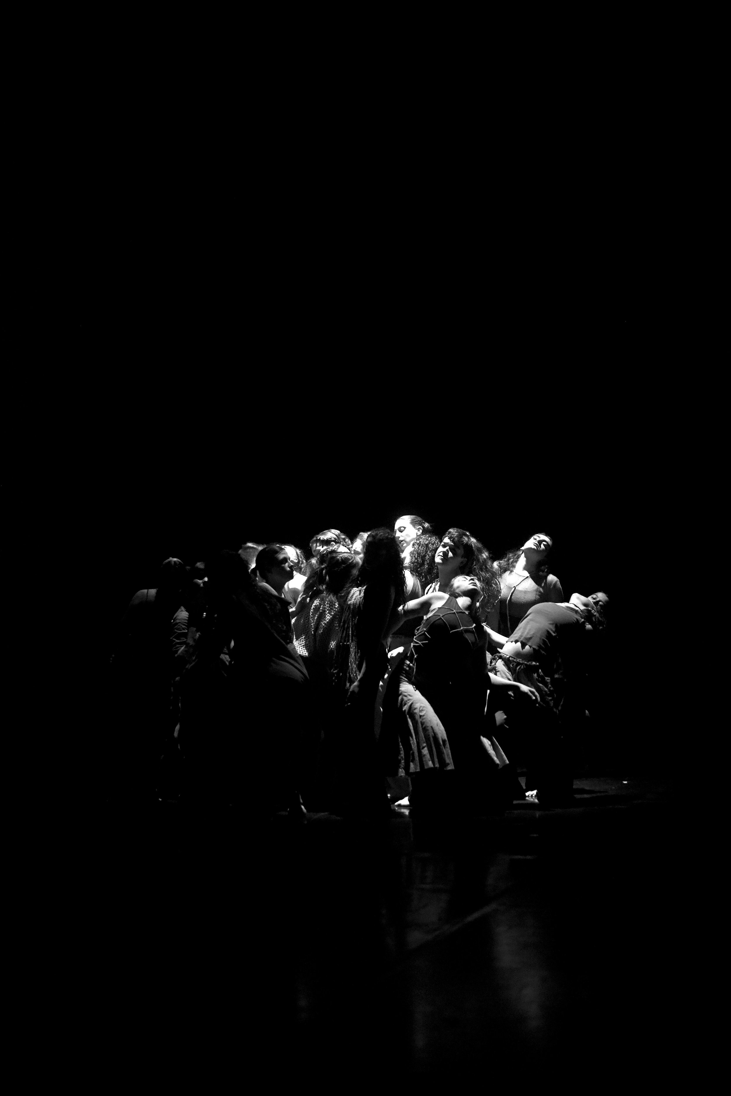Blick auf eine Gruppe von Menschen auf einer Bühne bei einer Theateraufführung, schwarz-weiß, Beleuchtung von unten, steht auf einer dunklen Bühne, drapiert und versammelt in einer Formation.