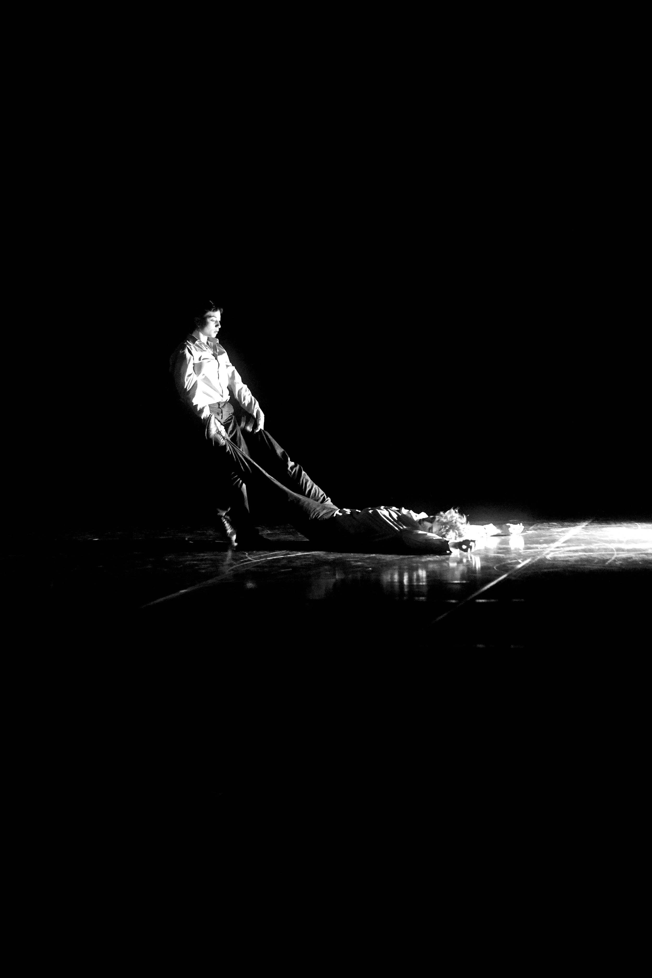 Schwarz-Weiß-Foto einer Person in einem weißen Hemd, die eine weitere Person auf dem Boden festhält. Die Szene spielt möglicherweise auf einer Bühne oder in einem Theater. Die Beleuchtung hebt die Personen hervor, während der Hintergrund dunkel ist.