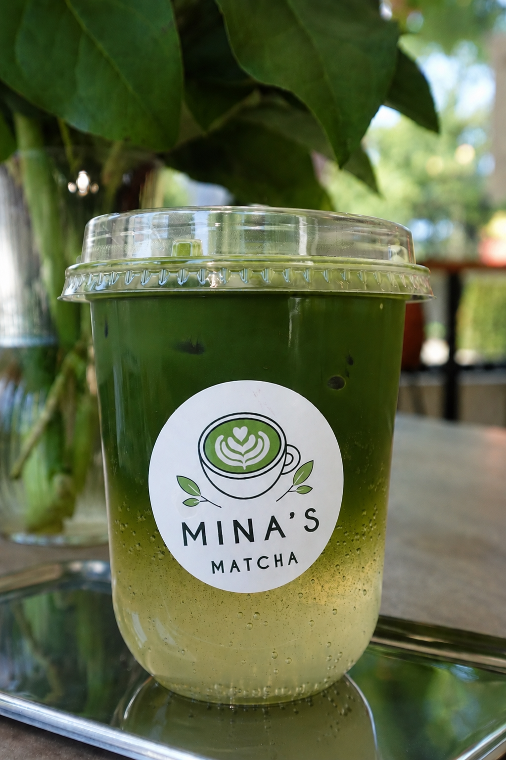 A plastic cup with a green and yellow matcha drink, labeled 'Mina's Matcha,' sitting on a silver tray. The background has green leaves and outdoor scenery with sunlight.