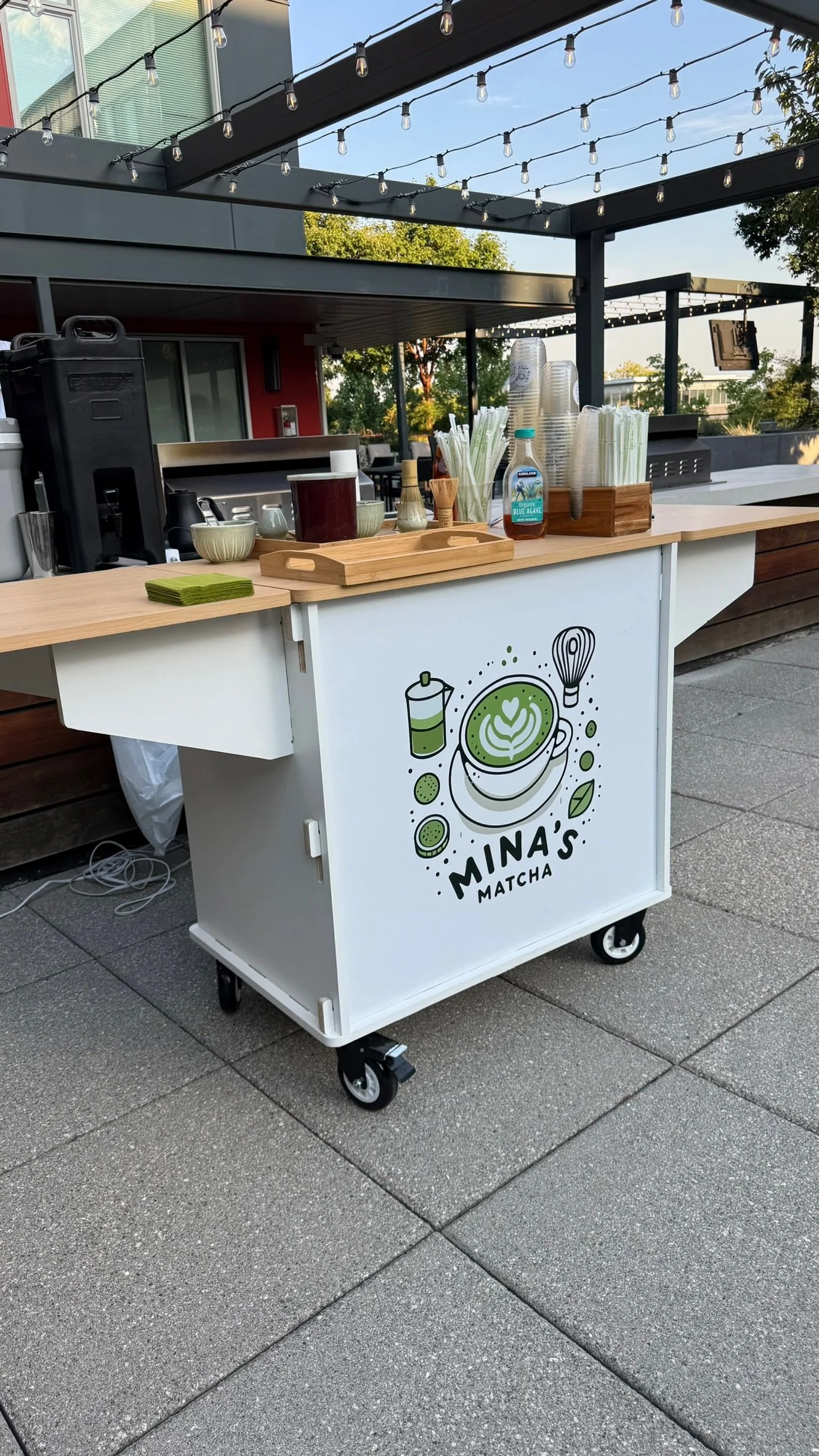 Mobile stand for Mina's Matcha, with matcha latte, green leaves, and matcha powder on the front. The stand has various utensils, cups, and a bottle of sweetener on top.