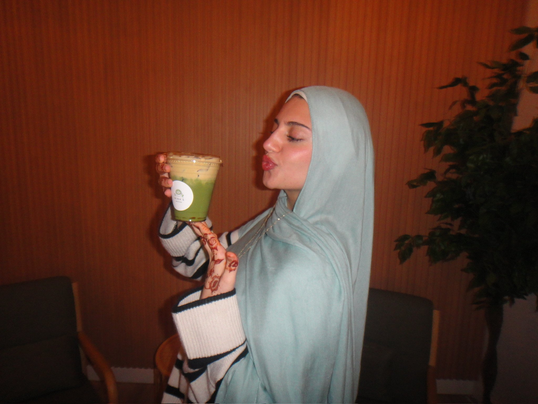 A woman wearing a light gray hijab with white and black striped sleeves is holding a large cup of matcha with cream top and some toppings, and making a kiss face toward the drink.